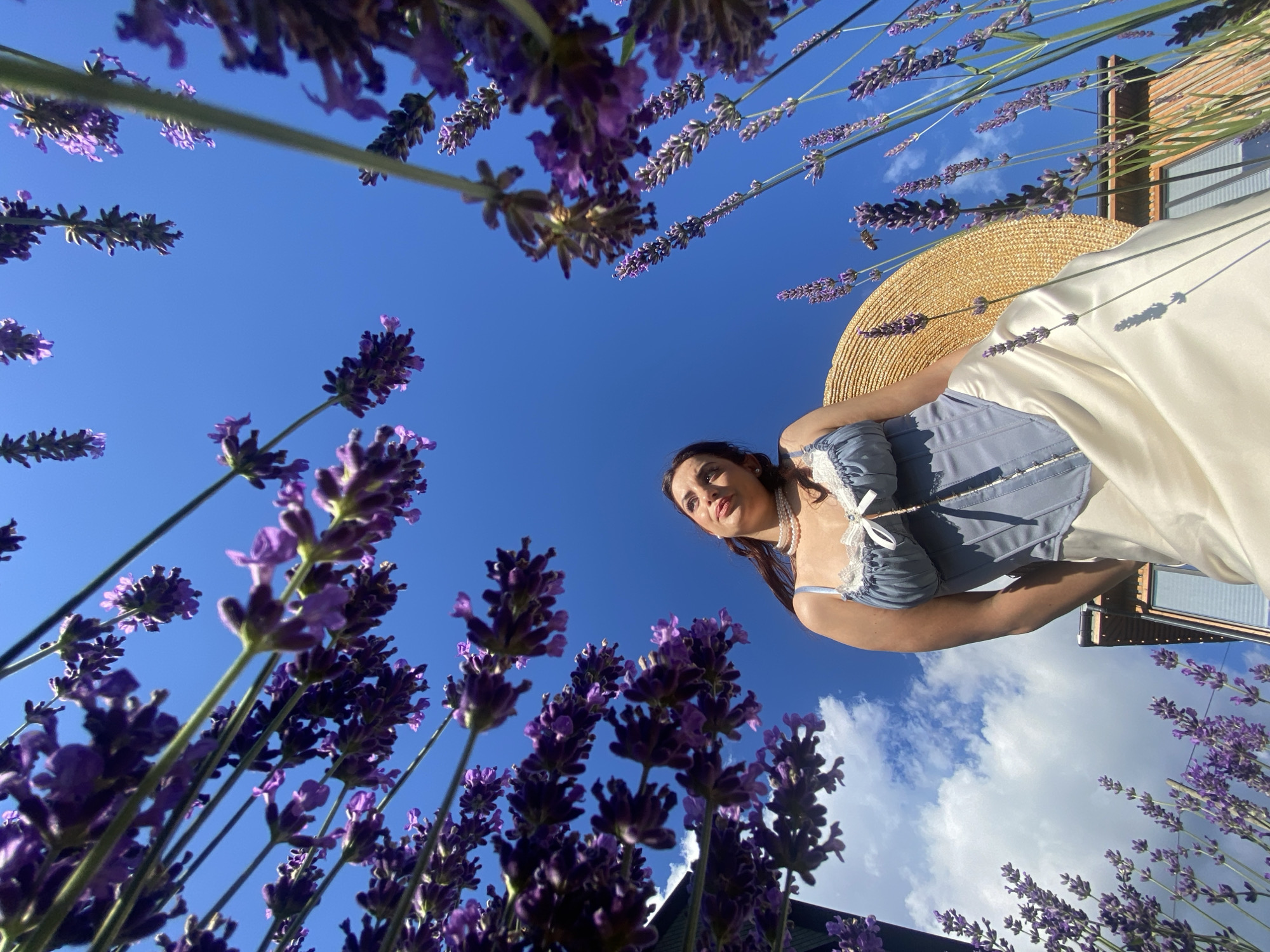 The image shows a low-angle shot of a young woman standing in a field of lavender under a bright blue sky. She is wearing a blue corset-style top with white lace trim and a white skirt. A straw hat is positioned behind her. The lavender plants are in full bloom, with clusters of purple flowers. The sky is clear and sunny, with a few white clouds visible. A building can be seen in the background, partially obscured by the lavender. The overall impression is one of a serene and idyllic outdoor scene. There is no information to suggest this location is Verkhneye Uchdere, Russia.