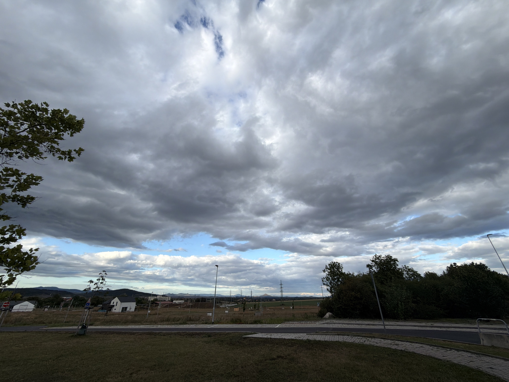 This is a daytime, wide-angle, outdoor photograph of a landscape in Louny, Czechia. The sky is filled with heavy, dramatic, gray clouds, with patches of blue sky and white clouds visible, particularly towards the horizon. In the foreground, there is a grassy area with a paved path. Beyond the path, there is a field with some sparse vegetation and a few scattered trees and saplings. In the middle ground, there are several houses, including a prominent white modern house with a dark roof, and a white building with a sloping roof. Further in the distance, there are more houses and buildings, and what appear to be power lines and transmission towers. The background consists of rolling hills and mountains. The overall impression is of a partly cloudy day in a semi-rural or suburban area.