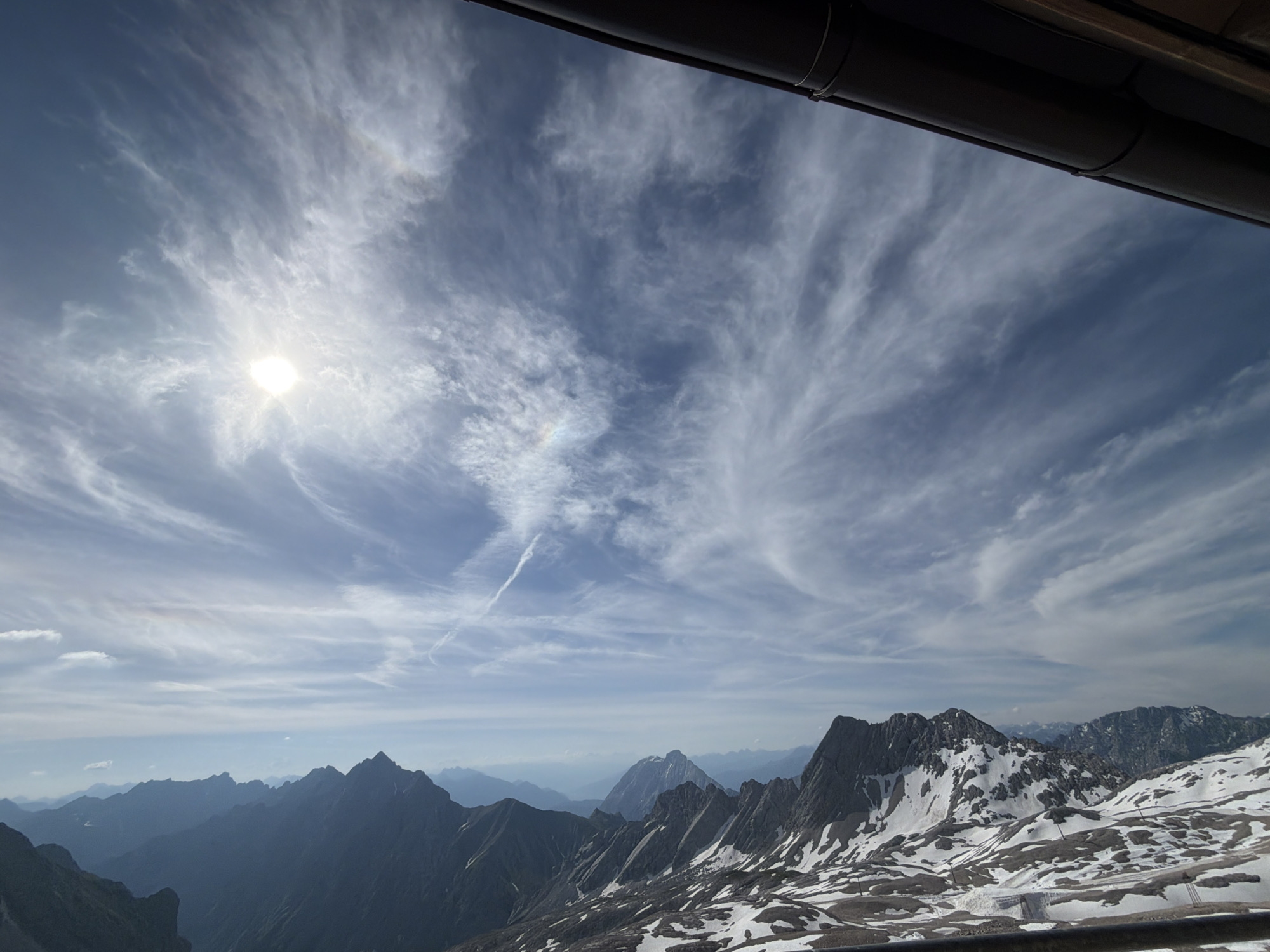 The image depicts a panoramic view of a mountainous landscape under a vast sky filled with wispy clouds. In the foreground, dark, shadowed mountain ridges with patches of snow are visible. The sun is brightly shining in the upper left portion of the sky, casting a lens flare. A faint rainbow-like halo or atmospheric optical phenomenon is present around the sun. Further into the distance, more mountain ranges fade into a hazy blue. The scene is framed by a dark, horizontal structure at the very top, possibly part of a building or viewing platform. The image is taken from a high vantage point, suggesting a location like the Zugspitze, Germany's highest peak, which is near Garmisch-Partenkirchen. There are no people or specific objects that indicate a city or urban environment. The scene is dominated by natural elements, with a focus on the dramatic interplay of light, clouds, and mountains.