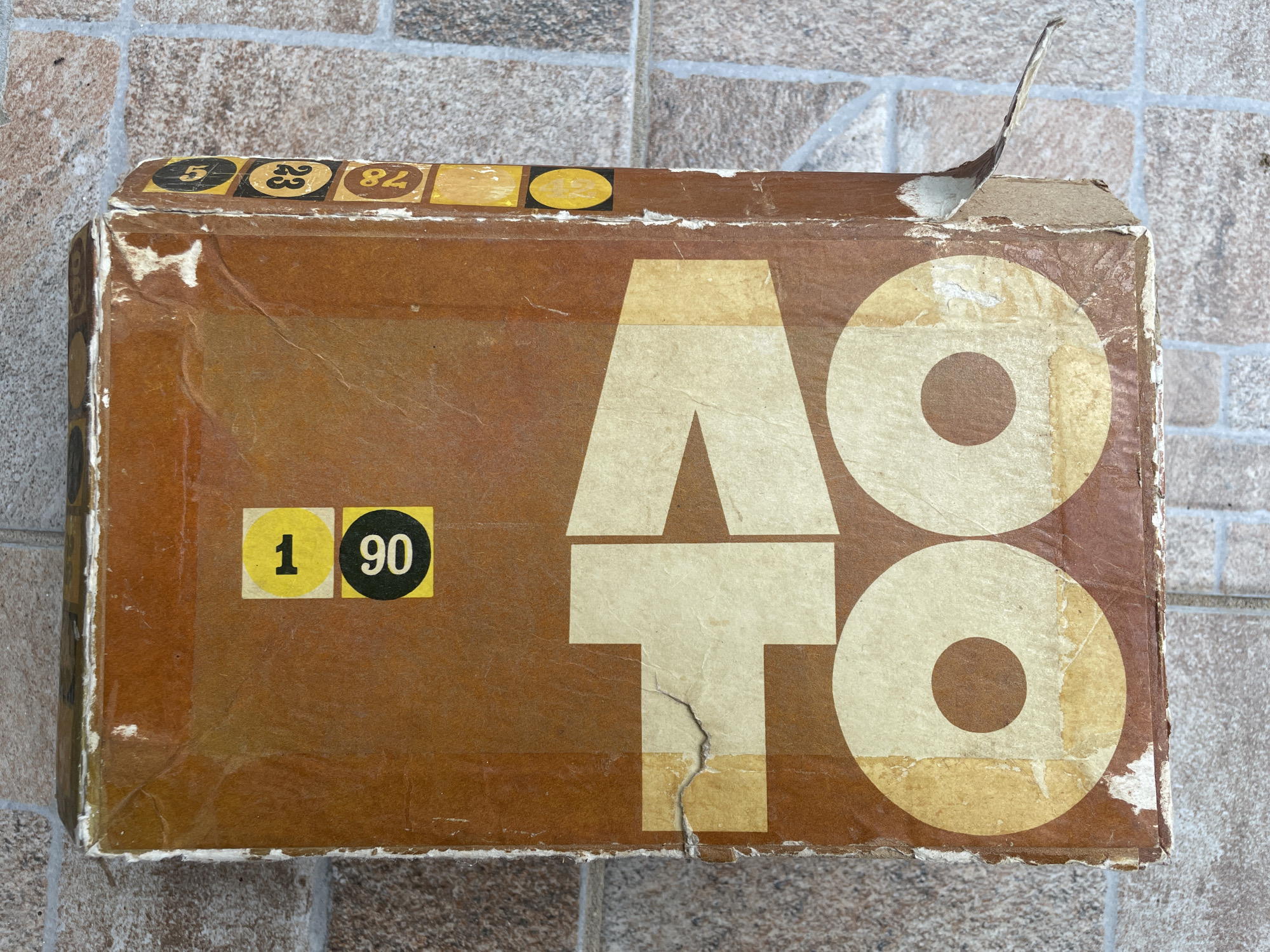 The image depicts a brown, aged cardboard box with the word "ATTO" printed on it in large, stylized white letters. The box appears to be worn, with frayed edges and areas where the surface is peeling or torn. In the top left corner, there are several small, round stickers with numbers printed on them: "5," "23," and "84," and another partly visible. On the left side, there is a yellow sticker with the number "1" and a black sticker with the number "90." A piece of the cardboard is peeling upwards on the upper right corner of the box. The box is placed on a tiled surface with a gray and brown patterned design. The scene is likely set in Byal Izvor, Bulgaria, although this is not explicitly evident from the image alone. There are no people visible in the image.