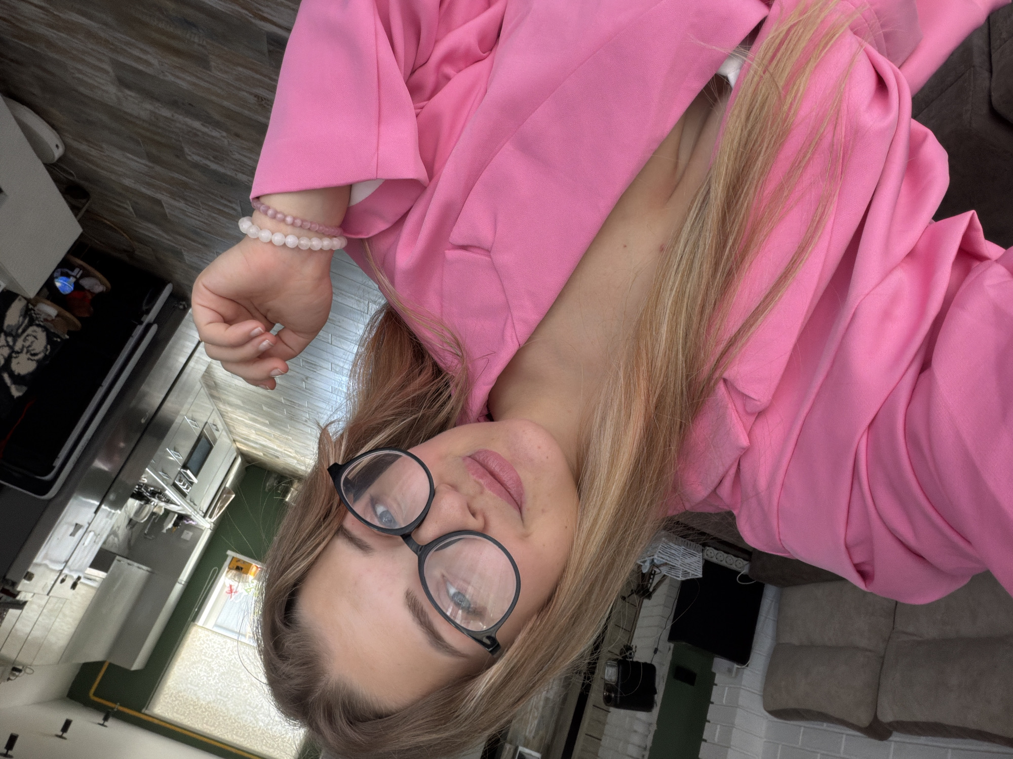 A selfie photograph shows a woman with blonde hair and glasses, wearing a pink blazer. She is indoors in what appears to be a home. In the background, a kitchen with stainless steel appliances is visible on one side, and a living room with a couch and television on the other side. The photograph is taken at an angle, likely held by the woman herself. There is no verifiable information to confirm the location is Большая Шильна, Russia.