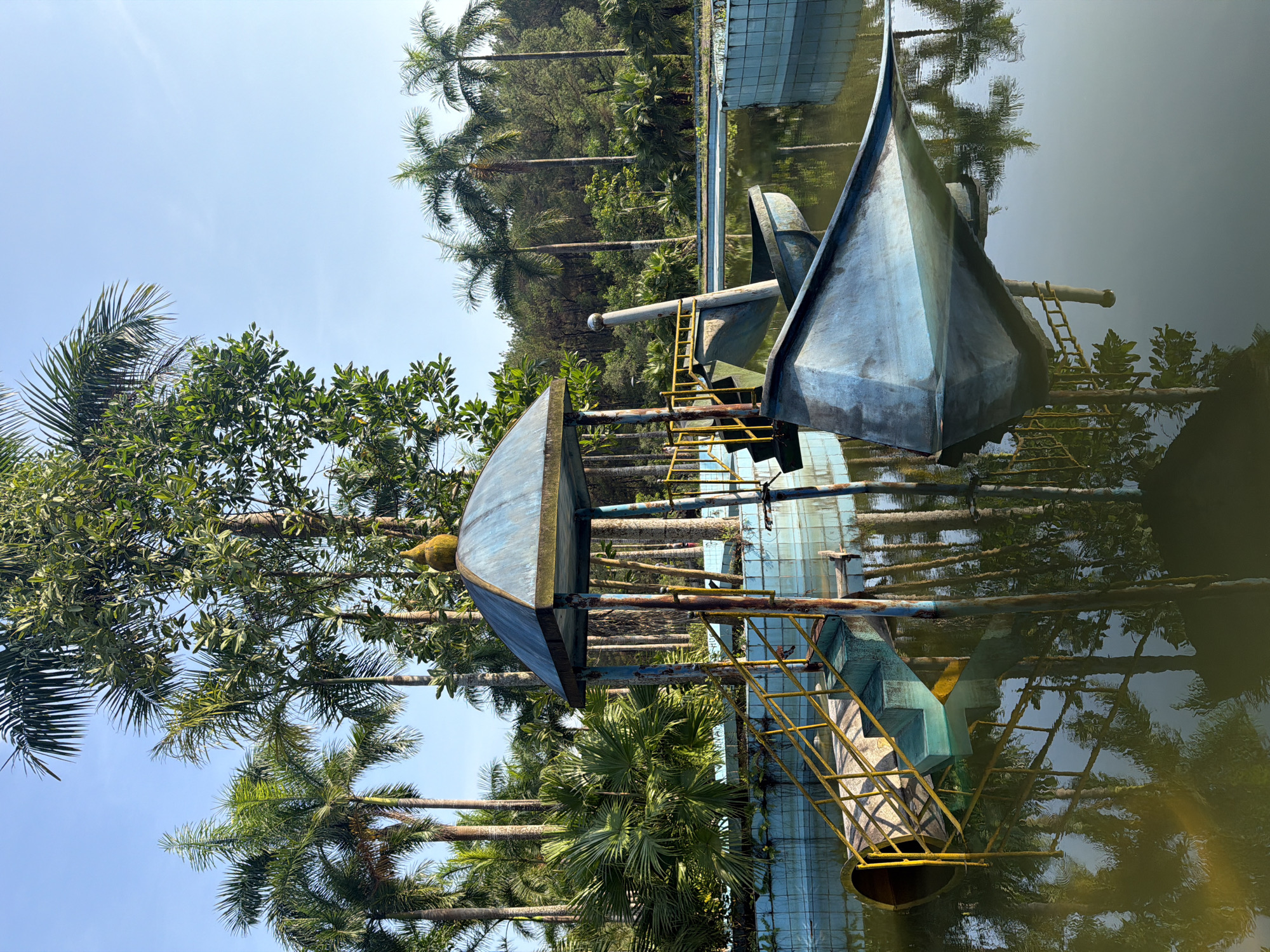 The image depicts a derelict boat-themed water structure, possibly a former water park feature, submerged in a stagnant pool. The structure, constructed from metal and concrete with remnants of blue paint, consists of a boat hull, a tower-like section, and connecting metal ladders. The pool is surrounded by palm trees and other vegetation, with their reflections visible in the murky water. The location is Thủy Xuân Ward, Vietnam.