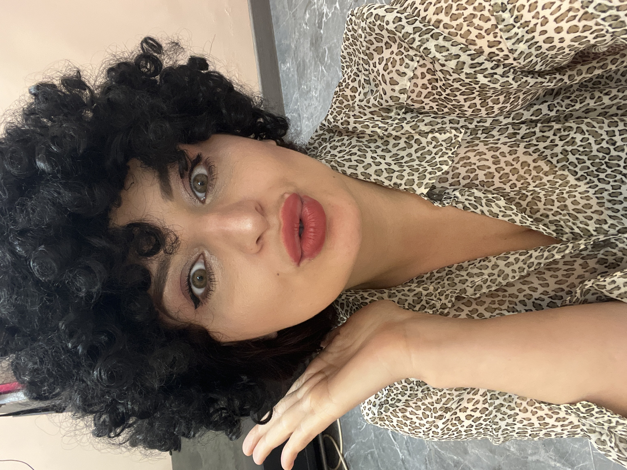 A front-facing, waist-up portrait captures a woman posing with a playful expression. She wears a voluminous, tightly curled black wig that frames her face and a sheer, light-colored leopard-print collared shirt. Her face is made up with prominent black winged eyeliner, light eyeshadow, and dark mascara. Her full lips are painted a matte red/coral and pursed into a "kissing" or "duck face." Her light brown eyes are wide open, directly engaging the viewer. Her left hand is raised beside her face, palm open and fingers slightly splayed, appearing to gesture or listen. The background is an indoor setting with a plain, light beige wall behind her upper body on the right and a grey, mottled floor visible to her left and below. Blurred dark objects and a white electrical cable are partially visible on the floor in the far left of the frame. The location is in the city of Unknown, Unknown.