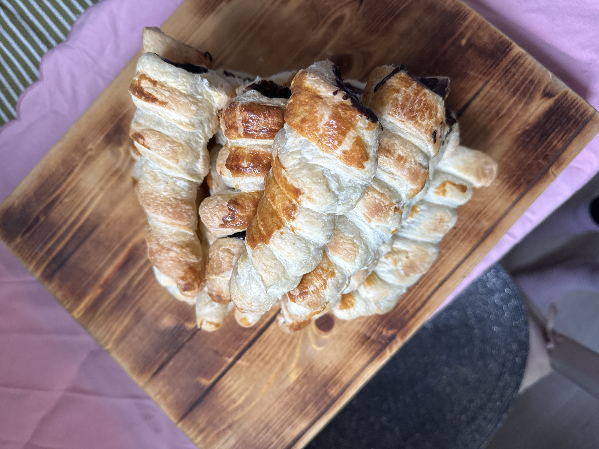 The scene presents approximately seven golden-brown puff pastries, each featuring a twisted or braided appearance and a dark, possibly chocolate, filling visible at the ends. The pastries are stacked on a rectangular wooden serving board with distinct grain patterns and darkened edges. This assembly rests upon a light pink fabric or surface. A pink scalloped-edge paper placemat partially occupies the upper right background, beneath which a green and white diagonally striped pattern is visible. A dark, textured, out-of-focus surface appears in the lower left. No identifiable features are present to determine a specific location within the city of Krasnoyarsk, Russia.