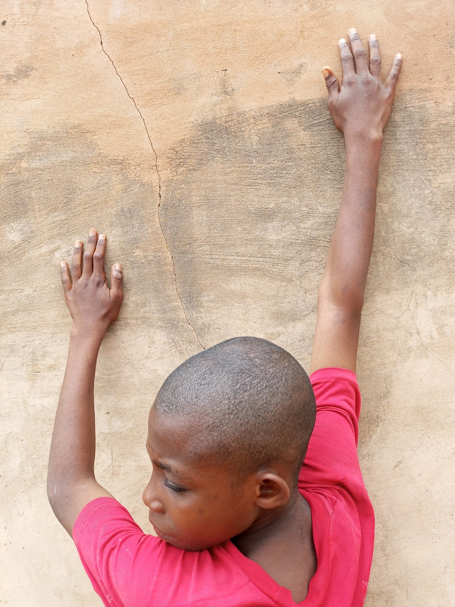 A close-up, high-angle shot shows a young Black person with a shaved head, wearing a bright pink t-shirt, pressing their hands against a textured, light brown wall that has a prominent vertical crack running through it. The person's face is partially visible, looking downwards and to the left. Their left arm is bent and raised, with their hand flat against the wall, fingers splayed. Their right arm is extended upwards along the wall, with their hand also flat and fingers spread. The lighting suggests an outdoor setting. The environment appears to be an exterior wall in Tempure, Nigeria.