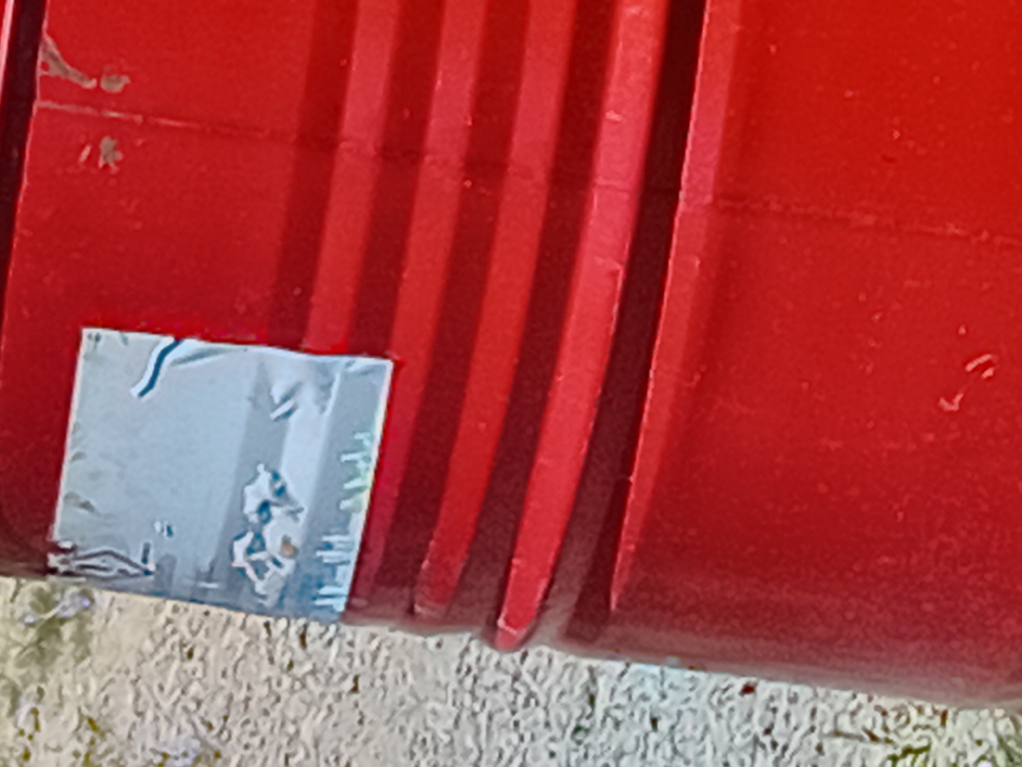 The image shows a close-up of a red, ribbed plastic barrel. A silver label with some blue markings is affixed to the upper portion of the barrel. The barrel is positioned outdoors on a surface that appears to be gravel or small stones, with some green vegetation visible in the background. The lighting suggests an outdoor, daytime scene. The location is specified as Verkhniachka, Ukraine.