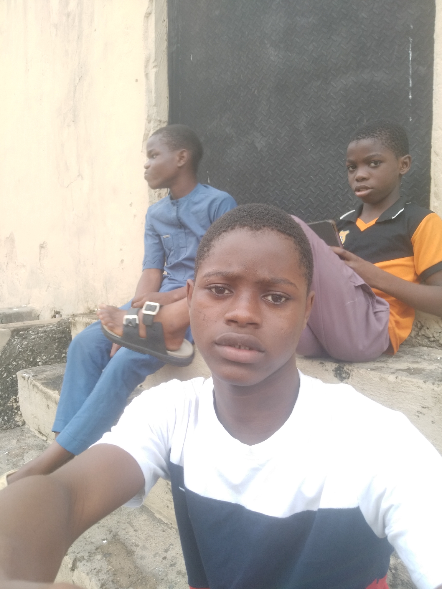 Three young African boys are sitting on concrete steps outside a building with a dark metal door. The boy in the foreground is taking a selfie, wearing a white and blue shirt. Behind him to the left, a boy in a blue shirt and sandals is looking to the side. To the right, another boy in an orange and black shirt is holding a phone. The building facade appears to be made of weathered concrete. The location is Unknown, Unknown.