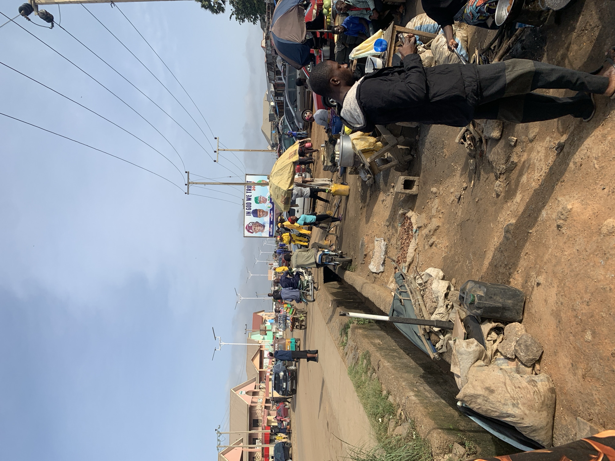 This media file captures a vibrant street scene in Rafin Par, Nigeria, under bright daylight with a clear blue sky. The image is oriented vertically (portrait mode), with the horizon running horizontally through the middle.
In the immediate foreground, a person wearing a black jacket and dark pants stands next to a makeshift stall, possibly engaged in food preparation, indicated by a metal pot and other items on a wooden stand. The ground is unpaved and dusty. A concrete drainage ditch runs alongside the road, filled with various debris including sacks, a black jerrycan, and pieces of wood.
The midground reveals a bustling street market environment. Several people are visible, some standing, others sitting or moving amongst an array of market stalls. Many stalls are covered by umbrellas or makeshift canopies, offering shade. Vehicles, including cars, motorcycles, and what appears to be a motorized tricycle (keke napep), are present on the road, suggesting a mix of parked and moving traffic. Along the left side of the road, several multi-story buildings with various architectural styles line the street. Modern streetlights are spaced along the road.
In the background, a large political billboard is prominently displayed on a utility pole. The billboard features multiple faces and the partially visible text "IN GOD WE TRUT" (likely "IN GOD WE TRUST") and "20__", suggesting an election campaign. Numerous power lines crisscross the sky, connected to utility poles. Shadows cast on the ground indicate it is likely midday or early afternoon.
The scene depicts a busy commercial area, characterized by roadside vendors, daily commerce, and local transportation, typical of an urban or semi-urban setting in Nigeria.