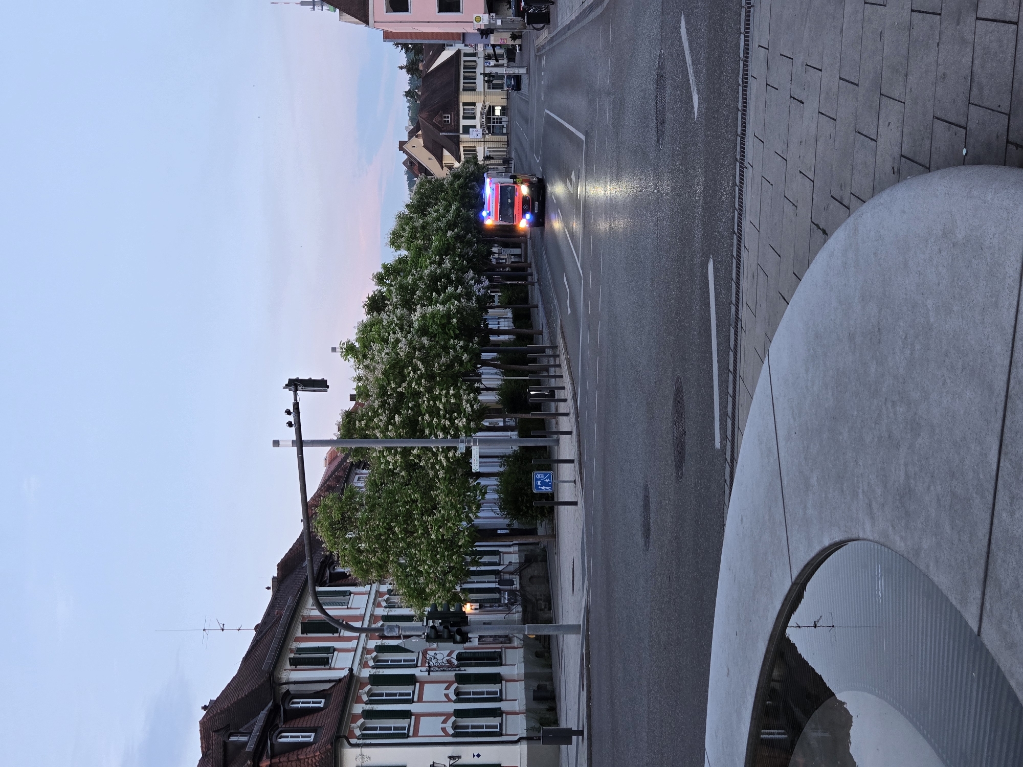 The image captures a street scene in Ansbach, Germany, near the Martin-Luther-Platz. In the foreground, a curved, concrete fountain edge is visible, with water reflecting the sky and surrounding structures. The street itself appears wet, possibly from recent rain, with lane markings and a manhole cover visible. On the left side of the street, traditional German buildings with red-tiled roofs, white facades, and green shutters line the pavement. Trees with white blossoms are planted along the sidewalk. A traffic light pole with traffic signals stands prominently on the left. In the distance, an emergency vehicle, likely an ambulance or fire truck, with its blue and red lights illuminated, is traveling down the street. The sky is overcast with soft pastel colors, suggesting either early morning or late evening. Several other buildings, including shops with signage, are visible further down the street, along with parked cars and streetlights. The overall atmosphere is calm and quiet, with the emergency vehicle being the most dynamic element in the scene.