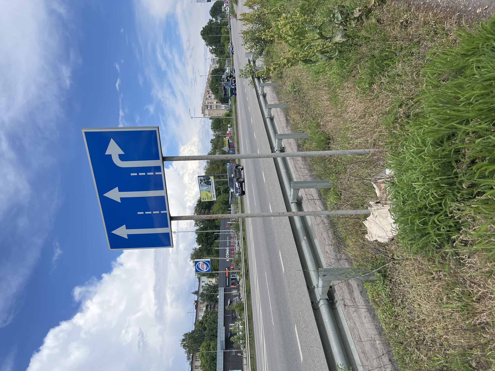 The image shows a road scene in Poltava, Ukraine. A blue road sign with white arrows indicates lane directions: two straight ahead and one turning right. The road is paved with gray asphalt and has white lane markings. A metal guardrail runs alongside the road, separating it from a grassy area. Buildings and trees are visible in the background. Cars are present on the road. The sky is blue with scattered clouds. A gas station is seen on the left side of the road.