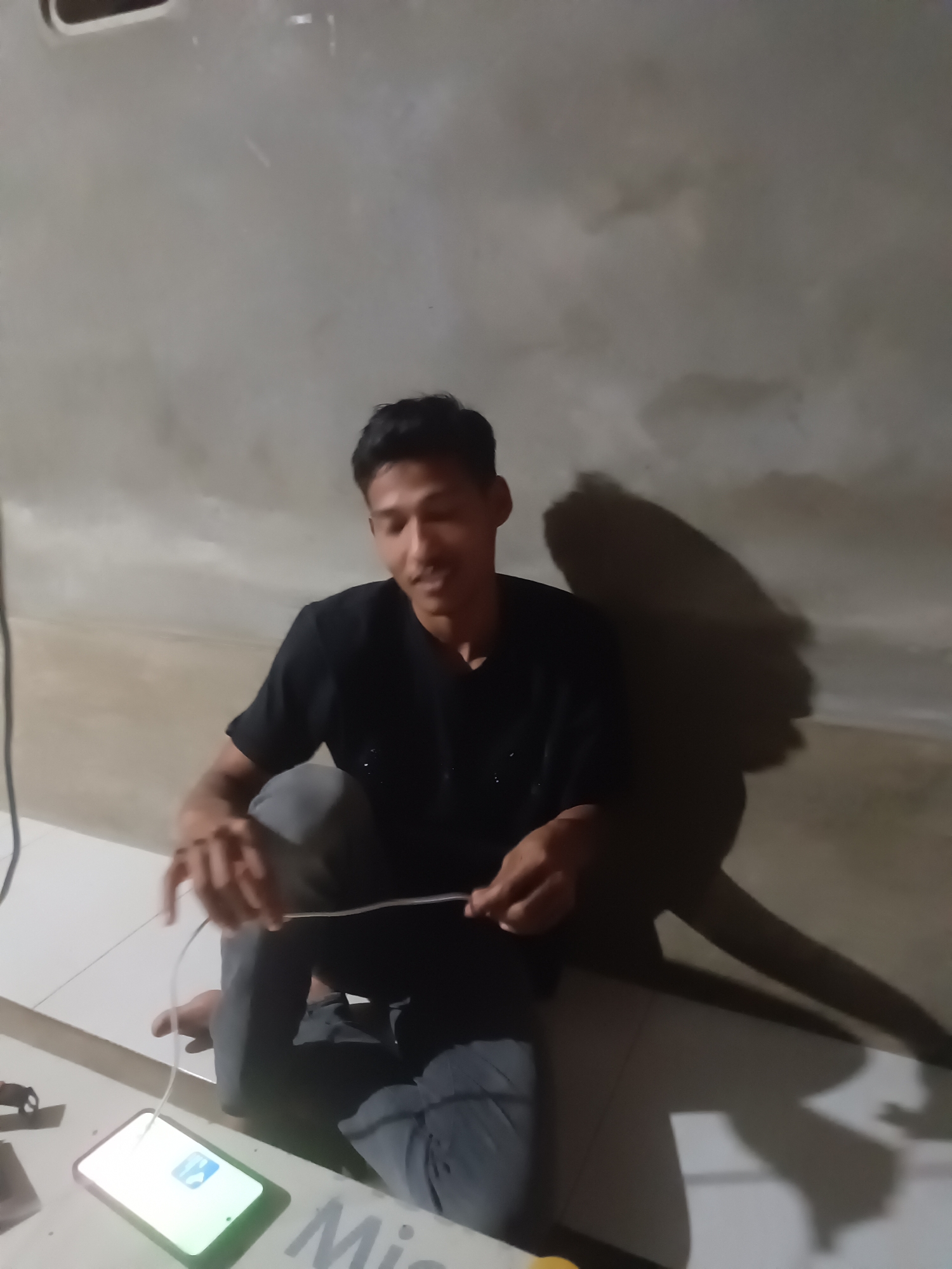 A young man wearing a black t-shirt and grey pants is sitting on the floor, holding a white cable that is connected to a smartphone on the ground. The smartphone displays a green icon. The man's face is turned slightly to his right, and he appears to be smiling. His shadow is cast on the wall behind him. The environment is a room with a concrete wall and tiled floor. The scene is illuminated by artificial light, creating distinct shadows. The image was taken in Lhokseumawe, Indonesia.