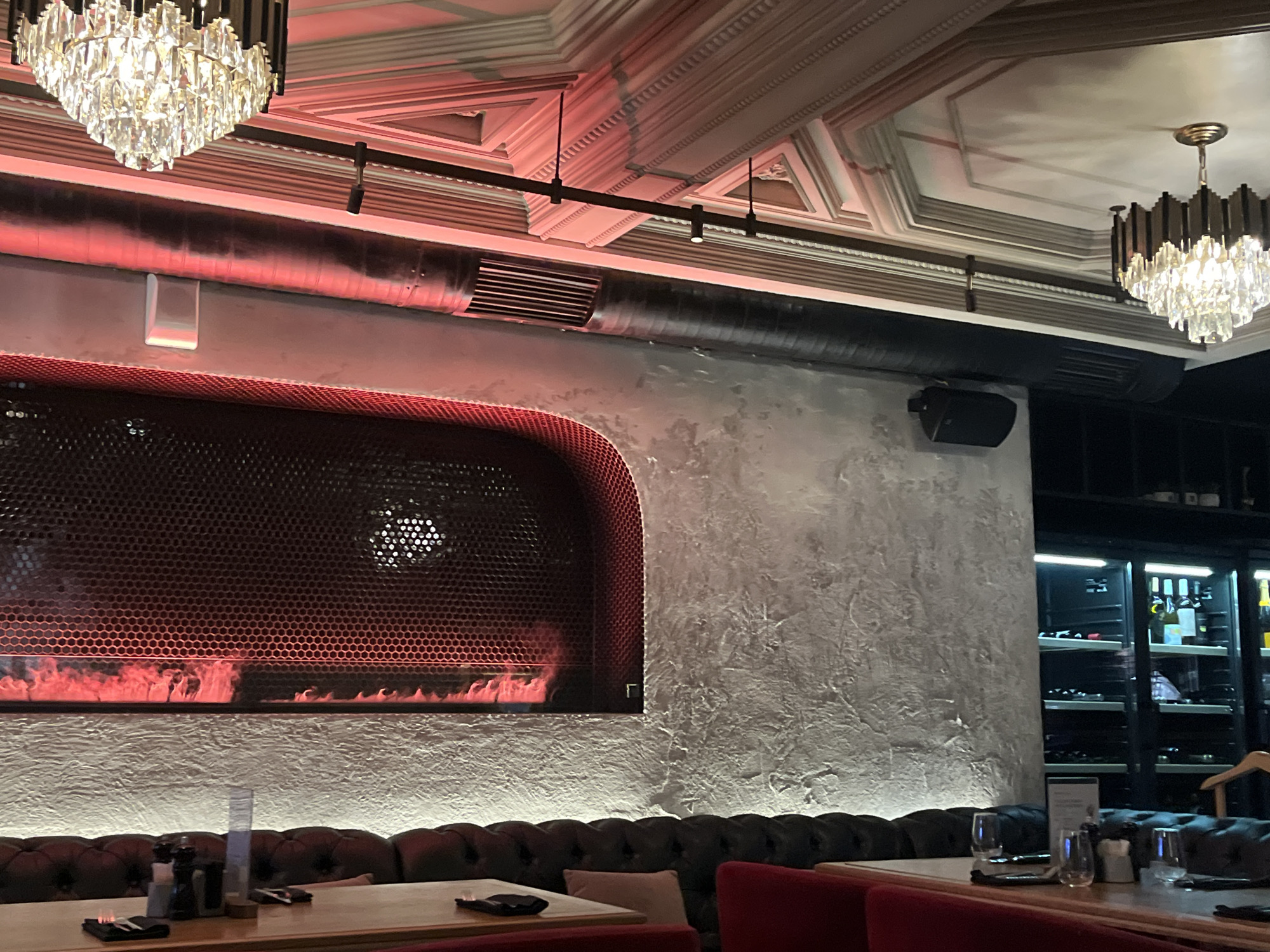 This image depicts the interior of a restaurant or bar, located in Unknown, Unknown. The scene is illuminated by a mix of ambient and accent lighting, creating a warm and contemporary atmosphere.
The ceiling features elaborate white classical molding and cornices, contrasted by exposed industrial-style black ventilation ducts and a black track lighting system with several small spotlights. Two prominent crystal chandeliers, one partially visible on the left and another on the right, hang from the ceiling, providing bright, sparkling light.
The upper left wall section has a dark, smooth surface with a strong reddish glow, likely from concealed red accent lighting positioned above a horizontal exposed pipe. Below this pipe, a white rectangular wall-mounted object is visible. The main feature on the left wall is a large, recessed, arch-shaped alcove with a dark hexagonal metal mesh screen. Behind this screen, a linear electric or decorative fireplace displays an orange-red flame effect. A circular vent with a similar mesh pattern is visible within the upper right portion of this alcove.
The wall to the right of the alcove has a textured, roughly rendered light grey or off-white surface. A black rectangular speaker is mounted on this wall.
Along the bottom of the image, a dark brown, tufted banquette provides seating, illuminated by a linear white light strip along its base. Wooden tables are set with place settings including black napkins, cutlery, various glasses (water, wine), and condiment shakers. Red upholstered chairs are partially visible at the very bottom right.
On the far right, a tall, dark display cabinet with glass doors and internal illumination showcases numerous bottles, suggesting a wine cooler or bar display. No people are visible in the scene.