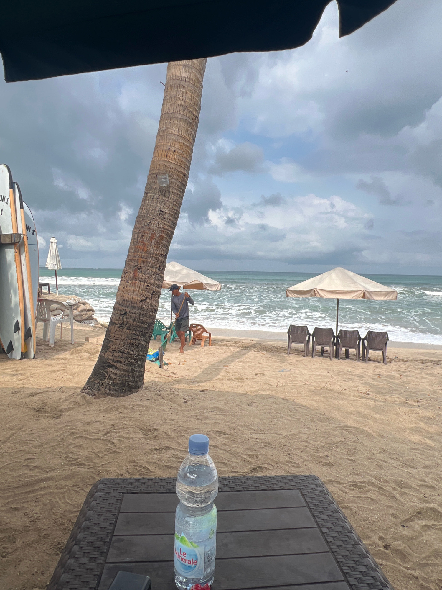 A wide shot captures a beach scene in Kuta, Indonesia, under a cloudy sky. In the foreground, a dark rattan table holds a partially full bottle of "Le Minerale" water and a black object. The sand is light brown with some debris.
Dominating the mid-ground is a large palm tree trunk leaning slightly to the left. To its left, a row of surfboards is propped upright. Further back, several beach chairs and a large white umbrella are set up on the sand, along with sandbags. A man wearing a dark shirt, shorts, and a cap is seen raking the sand with a long-handled tool.
The ocean stretches across the background, with moderate waves breaking near the shore. Several beach umbrellas and chairs are visible in the distance, suggesting a resort or public beach area. The sky is filled with heavy, gray clouds, obscuring any direct sunlight. The overall impression is a somewhat overcast day at a tropical beach.