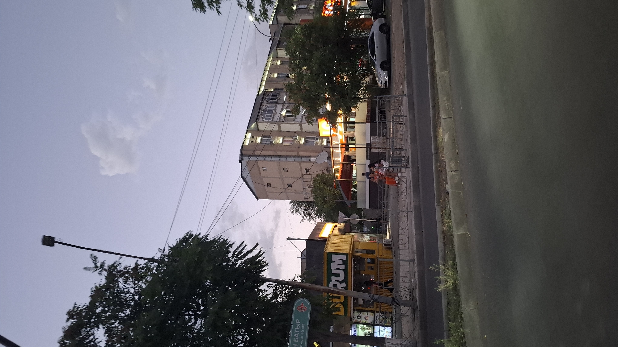 The image captures a street scene in Shymkent, Kazakhstan, likely during the late afternoon or early evening. In the foreground, a paved road with a curb is visible, transitioning into a sidewalk area. A yellow food kiosk with the word "DURUM" in large letters is a prominent feature on the left side of the scene. Adjacent to the kiosk, there's a green street sign with text in a foreign language and a decorative emblem.
Behind the kiosk, a multi-story building with a light-colored facade and numerous windows dominates the background. Several trees with dark green foliage are scattered throughout the scene, some in front of the building and others flanking the street. Another building, also multi-story and appearing to be a residential or commercial complex, is partially visible to the right of the first building. This second building has illuminated windows and awnings, suggesting it might house businesses.
Several individuals are present. Two people are seated on a bench near the food establishment, and another person is walking. A silver sedan is parked on the right side of the street, facing away from the camera. Overhead, power lines crisscross the sky, and a street lamp on a tall pole is visible on the left. The sky is a muted purple-gray, with a few scattered clouds, indicating a twilight setting. The overall impression is of a moderately busy urban environment with a mix of commercial activity and residential buildings.