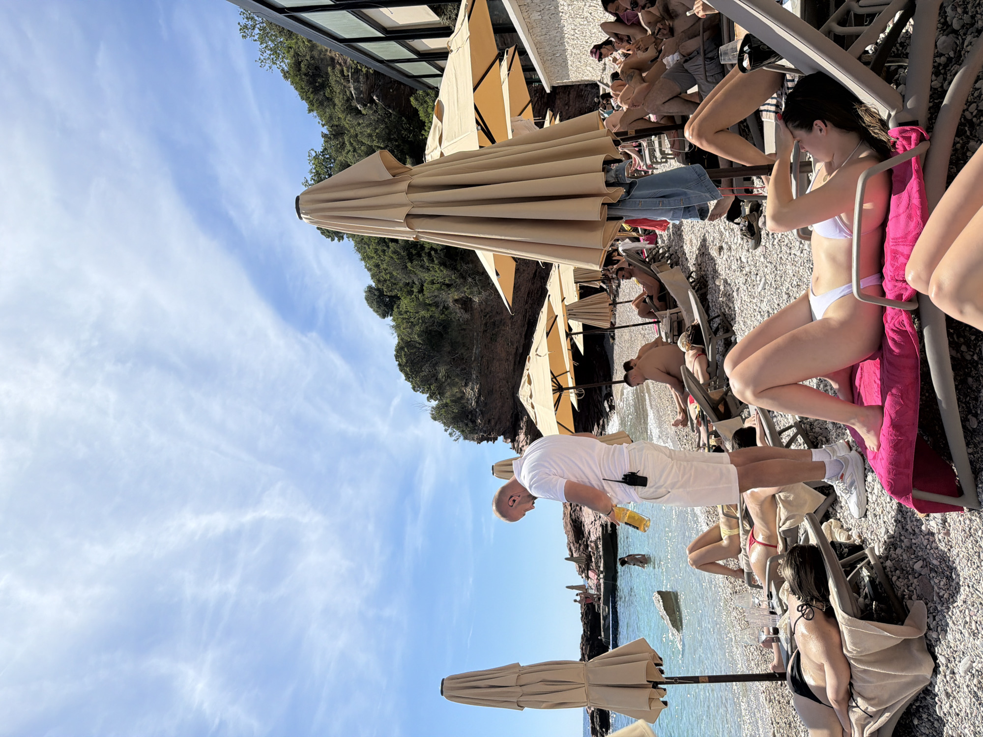 The image captures a beach scene at Sveti Stefan, Montenegro. A diverse group of people are on the beach, many lounging on beach chairs. Some are clad in swimwear. A few individuals are standing, one of whom is holding a drink. Beach umbrellas are interspersed amongst the beachgoers. The beach surface is pebbly. The sea is visible in the background. Buildings and vegetation are present in the upper portion of the picture. The sky is blue with clouds.
