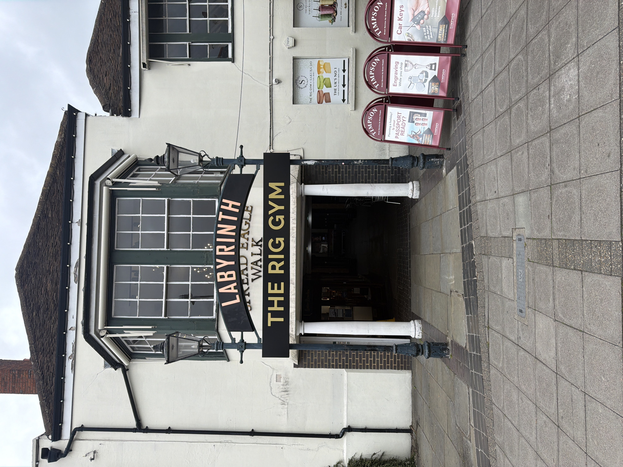 The image depicts a street scene in Epsom and Ewell, United Kingdom, focusing on the entrance to "The Rig Gym," located in the Labyrinth, off the Academic Eagle Walk. The entrance is marked by a dark sign with gold lettering. Above the entrance are windows with a dark green frame. To the right of the entrance, there are three Timpson signs advertising passport photos, engraving services, and car keys. The street is paved with grey and brown bricks. The overall lighting suggests an overcast day.