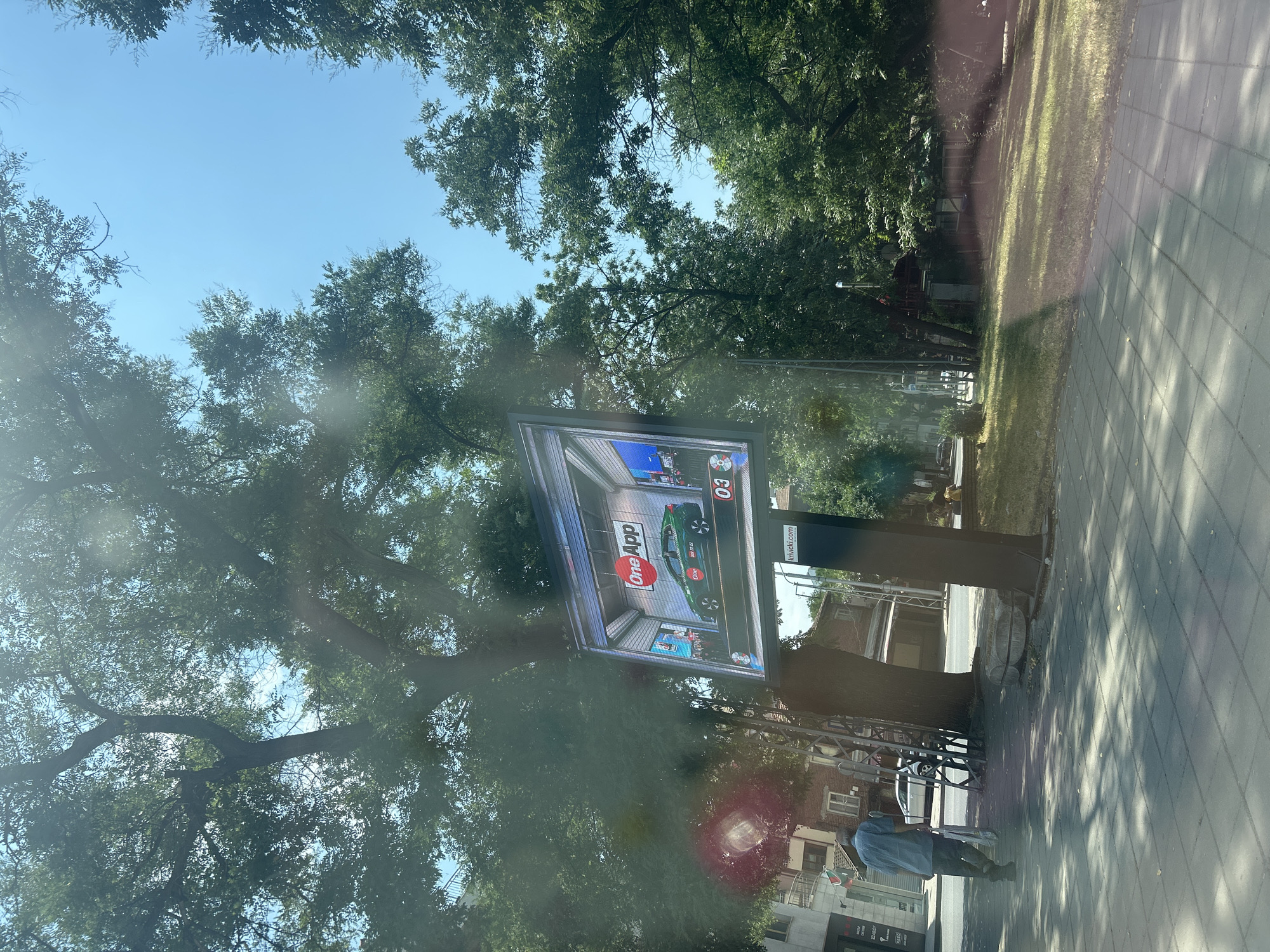 The image depicts an outdoor scene with a digital billboard as the central object, framed by lush green trees and a bright blue sky. The billboard displays an advertisement featuring a green car in a modern garage setting. The text "One App" is prominently visible in red and white on the advertisement, along with the number "03". To the right of the billboard, a smaller sign shows the website address "knivicki.com".
In the foreground, to the right of the billboard, a person wearing a light blue shirt and dark pants is walking on a paved sidewalk. The sun is shining, casting shadows from the trees and creating a dappled effect on the ground, suggesting it is daytime, likely late morning or afternoon given the angle of the sun. The setting appears to be a park or a tree-lined street in Plovdiv, Bulgaria, as indicated by the mention of the city. The overall atmosphere is bright and sunny, with no apparent emotional cues beyond the ordinary activity of a person walking by. The background shows some buildings, indicating an urban or suburban environment.