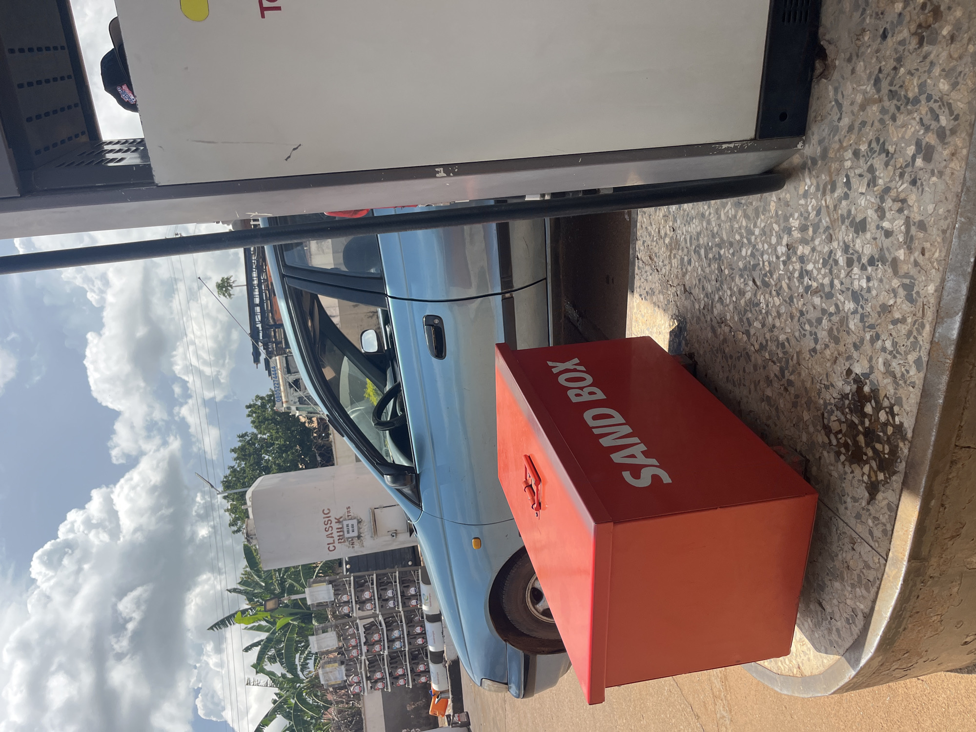 The image depicts a light blue sedan parked next to a gas pump at a station in Gura Top, Nigeria. In the foreground, a bright red sandbox with a red handle on top is positioned on a concrete circular base. The sandbox has "SAND BOX" written in white capital letters on its side. The car's driver's side door is open, and the interior is visible. Behind the car, a large white cylindrical tank labeled "CLASSIC BULK" and "REGO OIL NL 450" is situated, with rows of oil canisters stacked on shelves to its left. In the background, lush green trees and a bright blue sky with scattered clouds are visible. Power lines are also present in the distance. The ground is a mix of reddish-brown earth and speckled concrete. A person's hat, possibly a baseball cap, is visible on top of the gas pump.