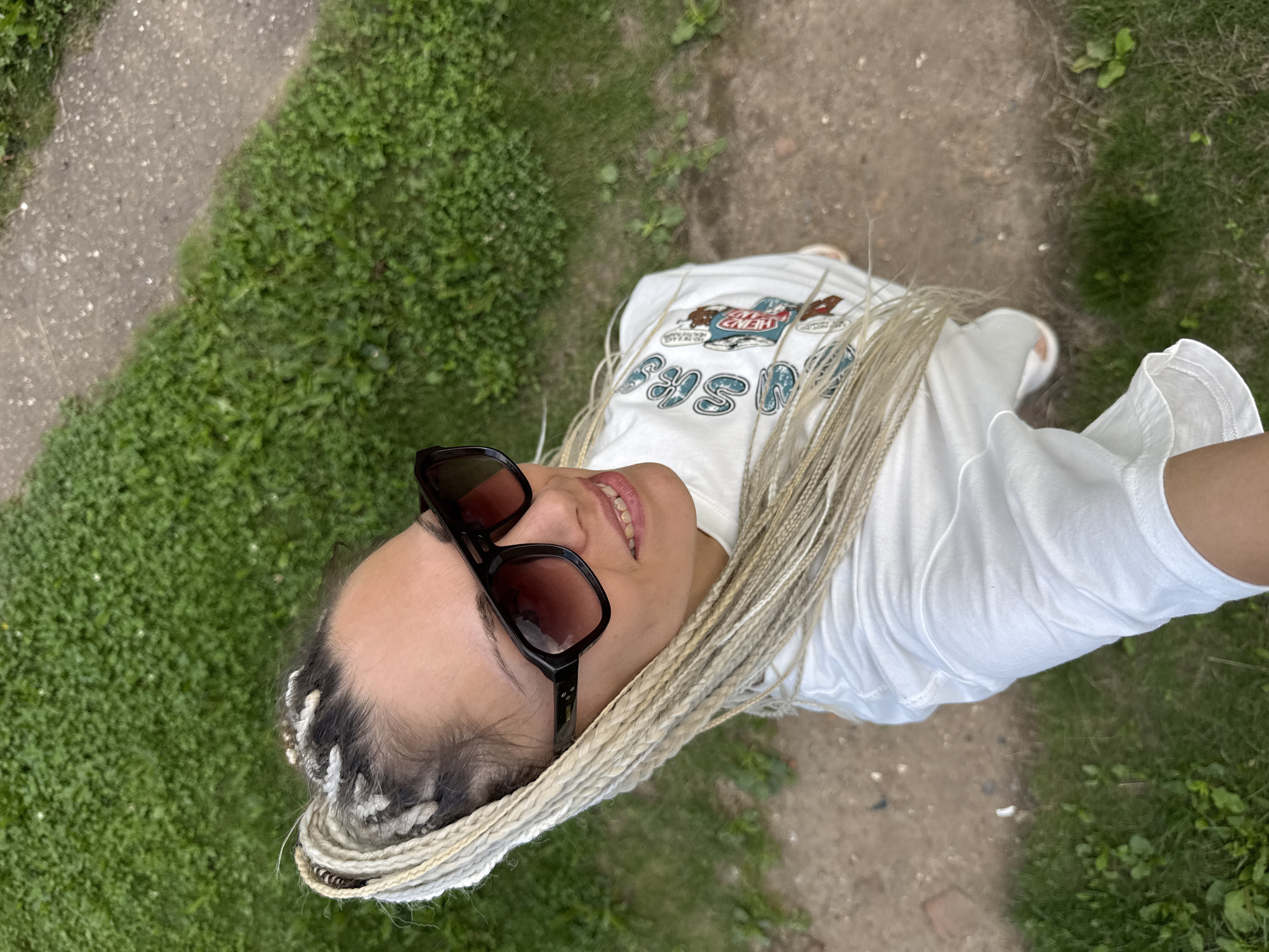 In the city of Rusanovo, Russia, a woman with light-colored braided hair and sunglasses is captured in a high-angle shot, taking a selfie. She is wearing a white t-shirt with a graphic print. The background consists of green grass and a dirt path. The overall scene is casual and outdoors.