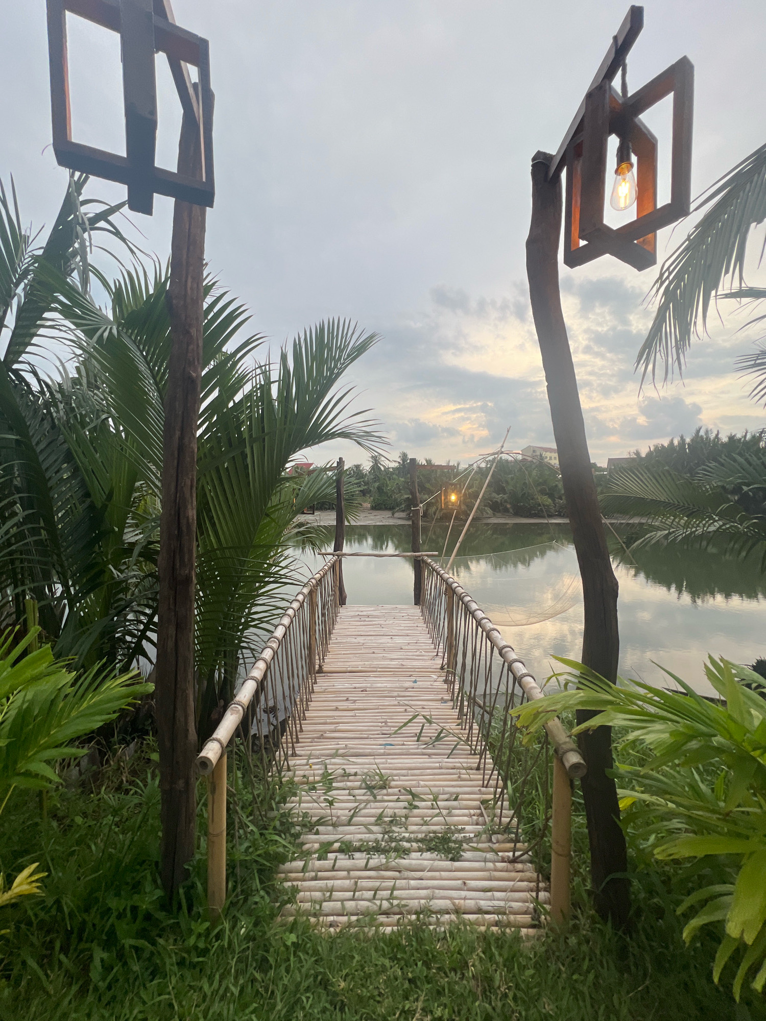 This is a medium-shot, eye-level photograph taken in Hoi An, Vietnam, at dusk. The image features a rustic bamboo walkway extending over a calm body of water, flanked by lush green palm trees and tropical foliage. Two wooden lampposts with angular, decorative frames stand on either side of the walkway, one of which has a lit Edison-style bulb. In the background, the water reflects the soft glow of the setting sun and the cloudy sky. A fishing net is visible suspended between poles on the water. The overall atmosphere is tranquil and natural, characteristic of the serene riverside landscapes found in Hoi An.
