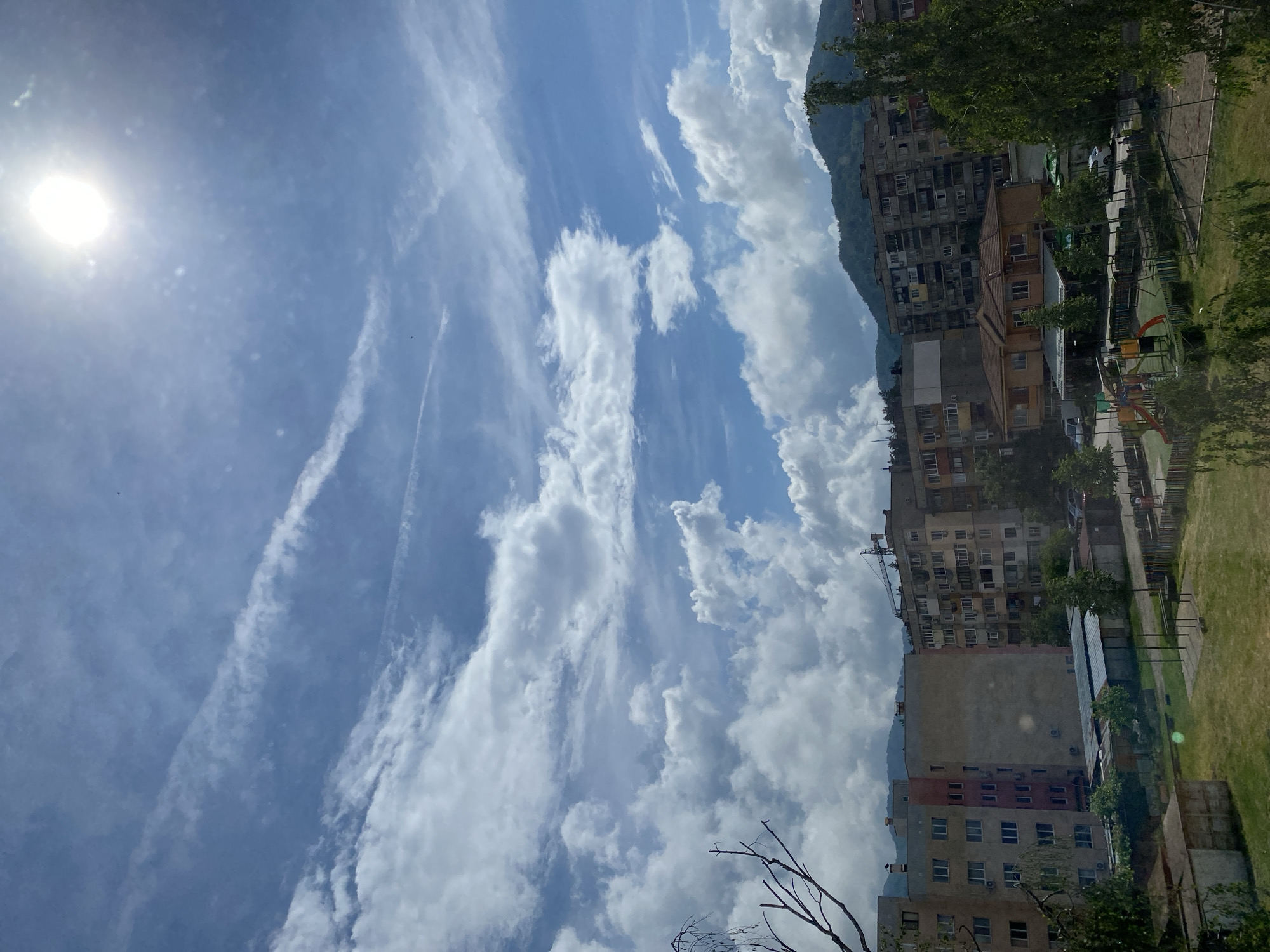 This image captures a vibrant, sunny day in Troyan, Bulgaria, showcasing a blend of urban architecture and natural scenery. The composition is split, with a dominant blue sky filling the upper and left portions, and residential buildings, green spaces, and mountains occupying the right and lower sections.
The sky is bright blue, illuminated by an intense, circular sun in the upper left corner, indicating midday or early afternoon. Scattered white cumulus clouds drift across the expanse, interspersed with wispy cirrus clouds and several faint contrails from aircraft.
The urban elements consist of multiple multi-story apartment buildings, characteristic of post-socialist architecture, with visible windows and balconies. These structures vary in color, ranging from light grays and browns to a distinct reddish section on one building. Behind some of these buildings, the dark, green-covered slopes of mountains are visible, suggesting a mountainous region. In the foreground and interspersed among the buildings, there are lush green grassy areas, mature trees, and a colorful children's playground with slides and climbing structures. Some bare tree branches are visible in the very bottom foreground. Faint glimpses of parked cars can be seen between the buildings and foliage.
No specific activities are taking place, and no people are visible in the frame. The scene conveys a sense of a peaceful, ordinary day in a residential area, framed by impressive natural surroundings. There is no visible text in the image.