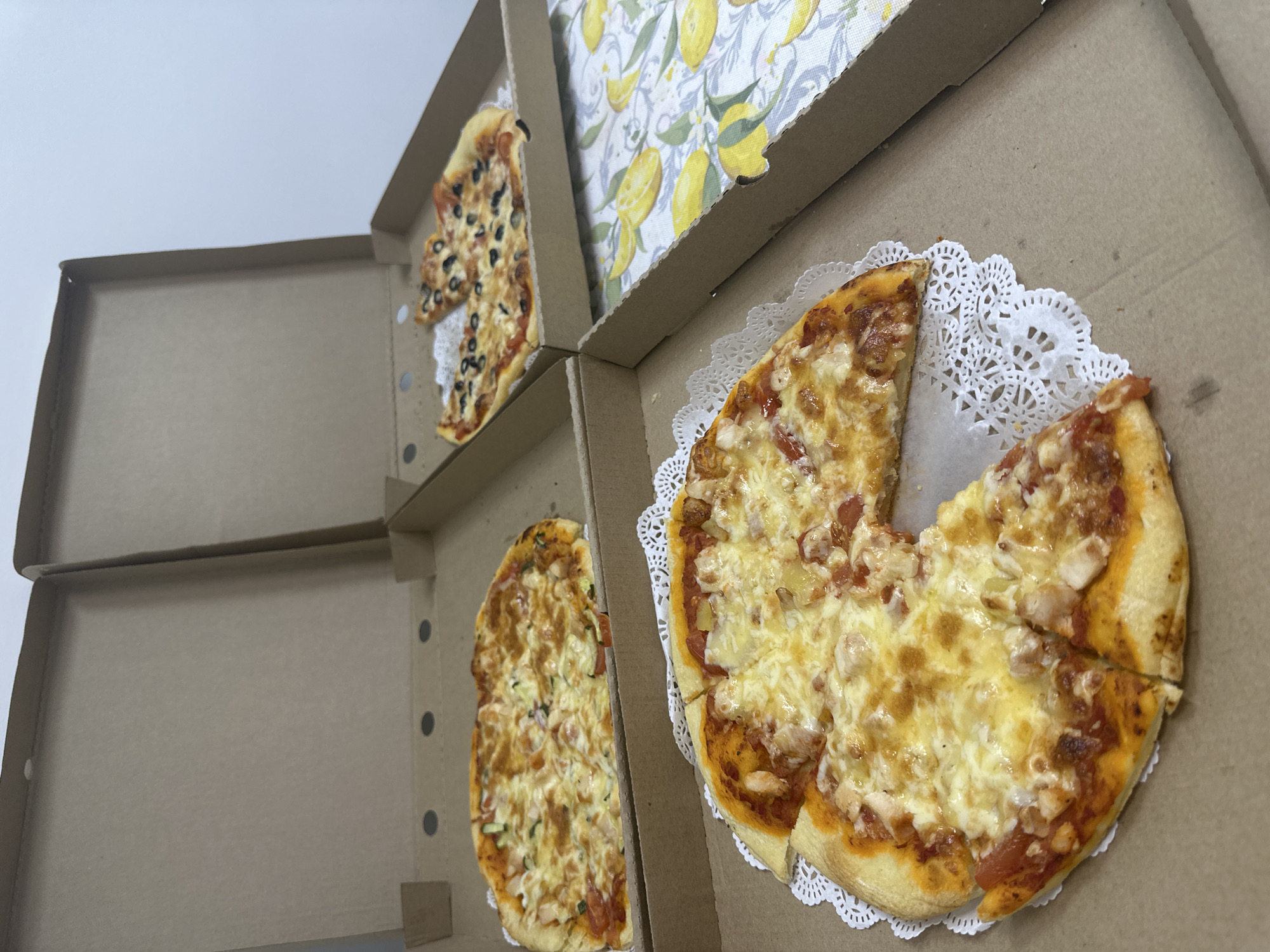 The image shows three partially eaten pizzas in cardboard pizza boxes. One pizza appears to have black olives as a topping. Another pizza is topped with what seems to be chicken and pineapple. The third pizza has visible zucchini and tomatoes. The pizzas are sitting on white lace doilies inside their boxes. A yellow lemon patterned lining is visible inside one of the pizza boxes. Based on the appearance of the boxes and the food, it is likely a takeout meal. There is no evidence to suggest this picture was taken in Chaikovsky, Russia.