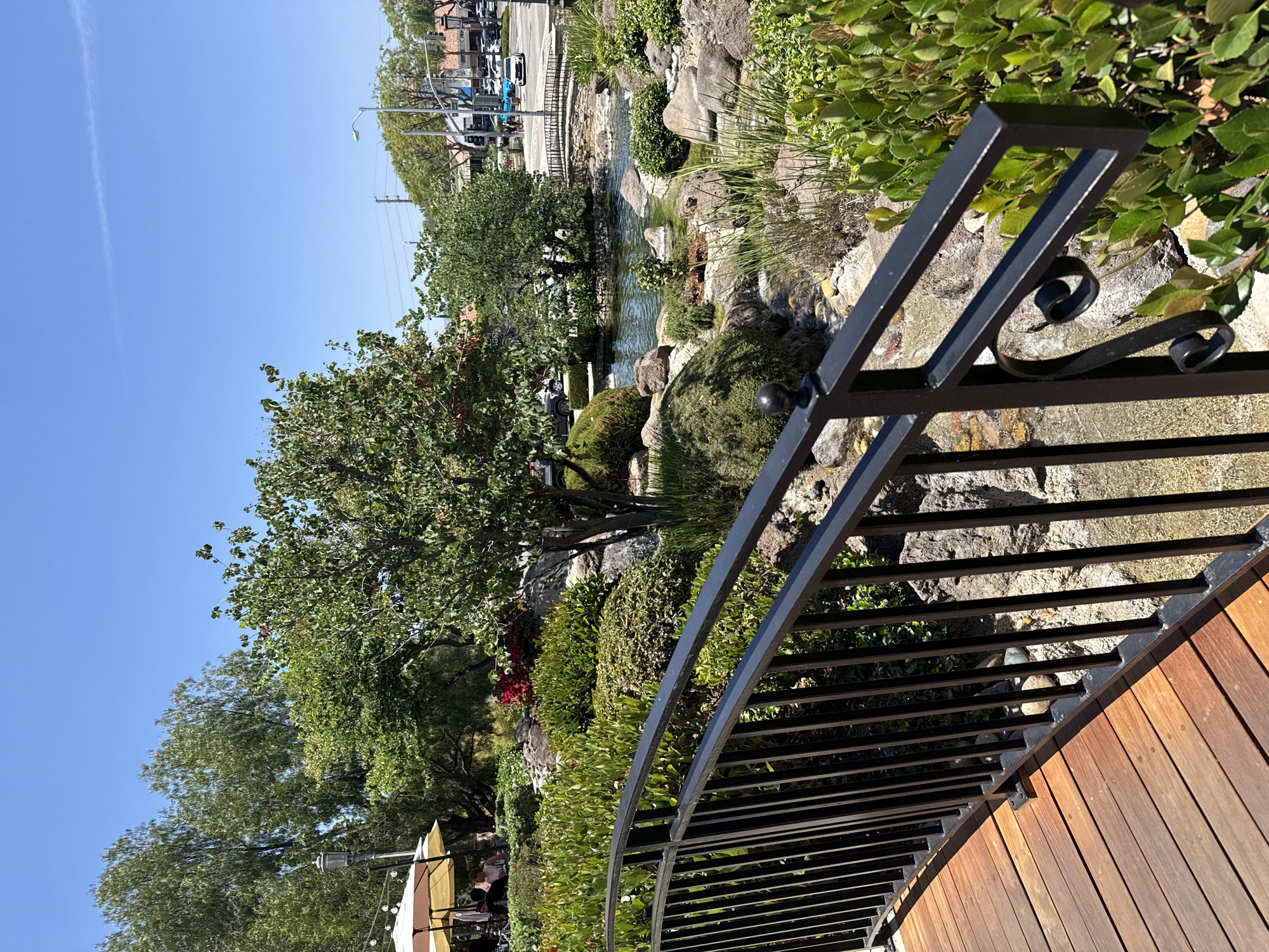 A medium shot captures a scenic view of a landscaped garden with a water feature in Calabasas, California, on a sunny day. In the foreground, a wooden deck with a black metal railing extends from the bottom left corner. Beyond the railing, a man-made stream cascades over rocks, feeding into a small pond. Lush green bushes and trees surround the water feature, with a larger tree in the center of the frame. To the left, an outdoor dining area with tables and umbrellas is visible, with a few people seated. In the background, a street with traffic, shops, and buildings is apparent, suggesting an urban or suburban setting. The sky is clear and blue with a faint white streak of a contrail.