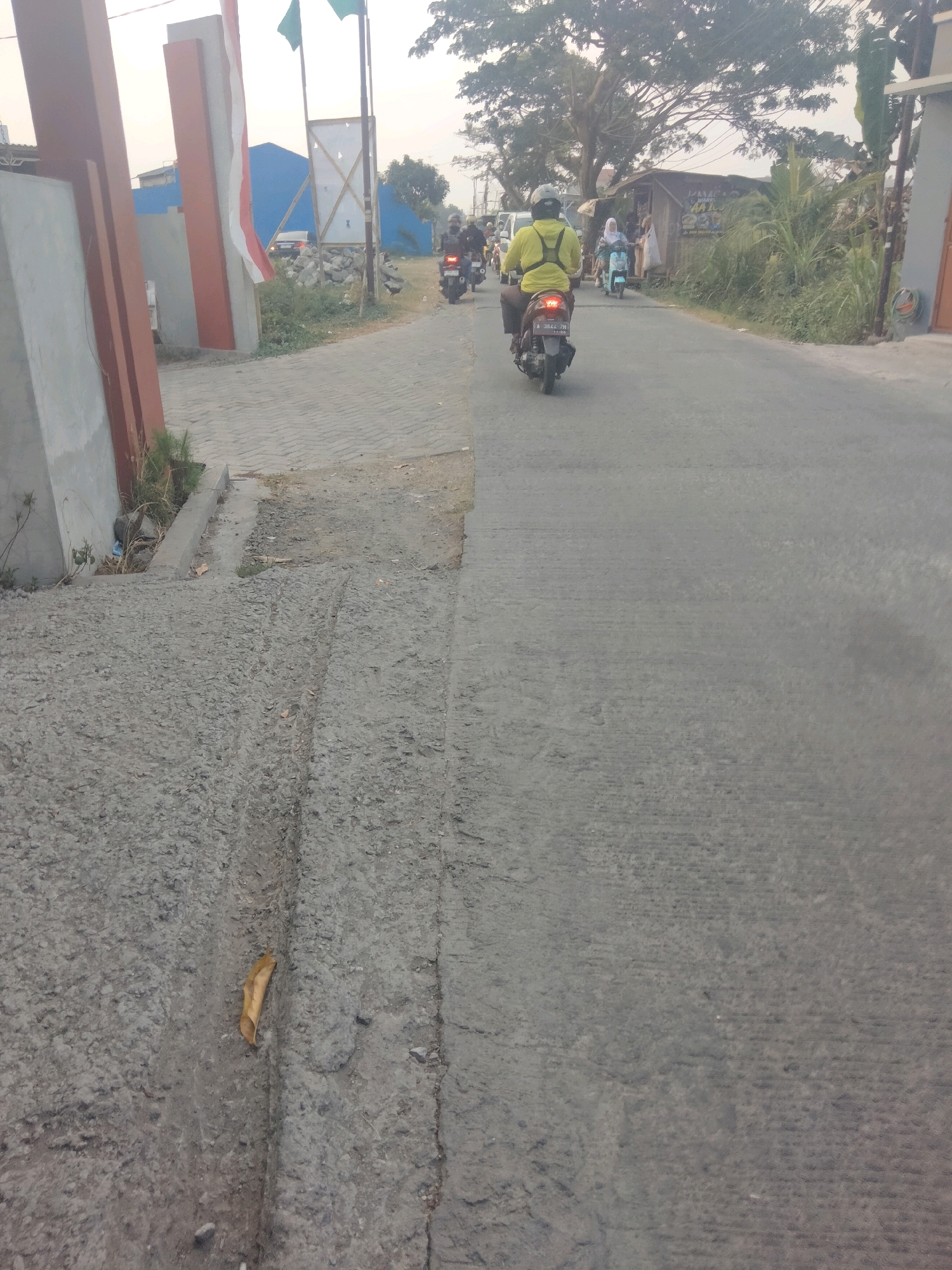The image depicts a street scene in Suka Sari, Indonesia. In the foreground, the rough, textured surface of a paved road is visible, with tire tracks impressed into it and a dry leaf lying in a crevice. To the left, a grey concrete wall with reddish-brown vertical beams and a white section with a red stripe is partially visible, suggesting the entrance to a building or property. Further down the road, several people are riding motorcycles. One person in a bright yellow jacket is riding a motorcycle towards the viewer. Another person on a lighter-colored motorcycle is ahead of them, and further in the distance, more motorcycles and pedestrians can be seen. The road itself is asphalt, with a lighter-colored paved shoulder on the left. Greenery, including trees and bushes, lines the right side of the road. A small wooden stall or building is also visible on the right, with signs in Indonesian. Two green flags are flying on poles in the background, and a blue tarp covers a structure. The sky is overcast.