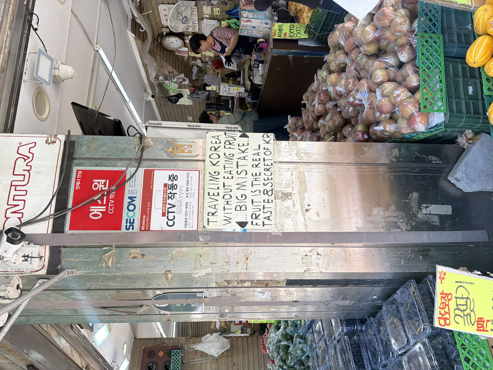 The image shows an outdoor market stall in Seoul, South Korea. A sign reads: "TRAVELING KOREA WITHOUT EATING FRUIT... BIG MISTAKE. FRUIT IS THE REAL TASTE & SECRET OF KR." Several people are visible, including a woman in a striped shirt and black gloves working at the stall. Objects visible include a CCTV security system, Korean signage, stacks of apples in plastic bags, and melons. The stall is constructed with metal and wood, and the environment appears to be a bustling commercial area.