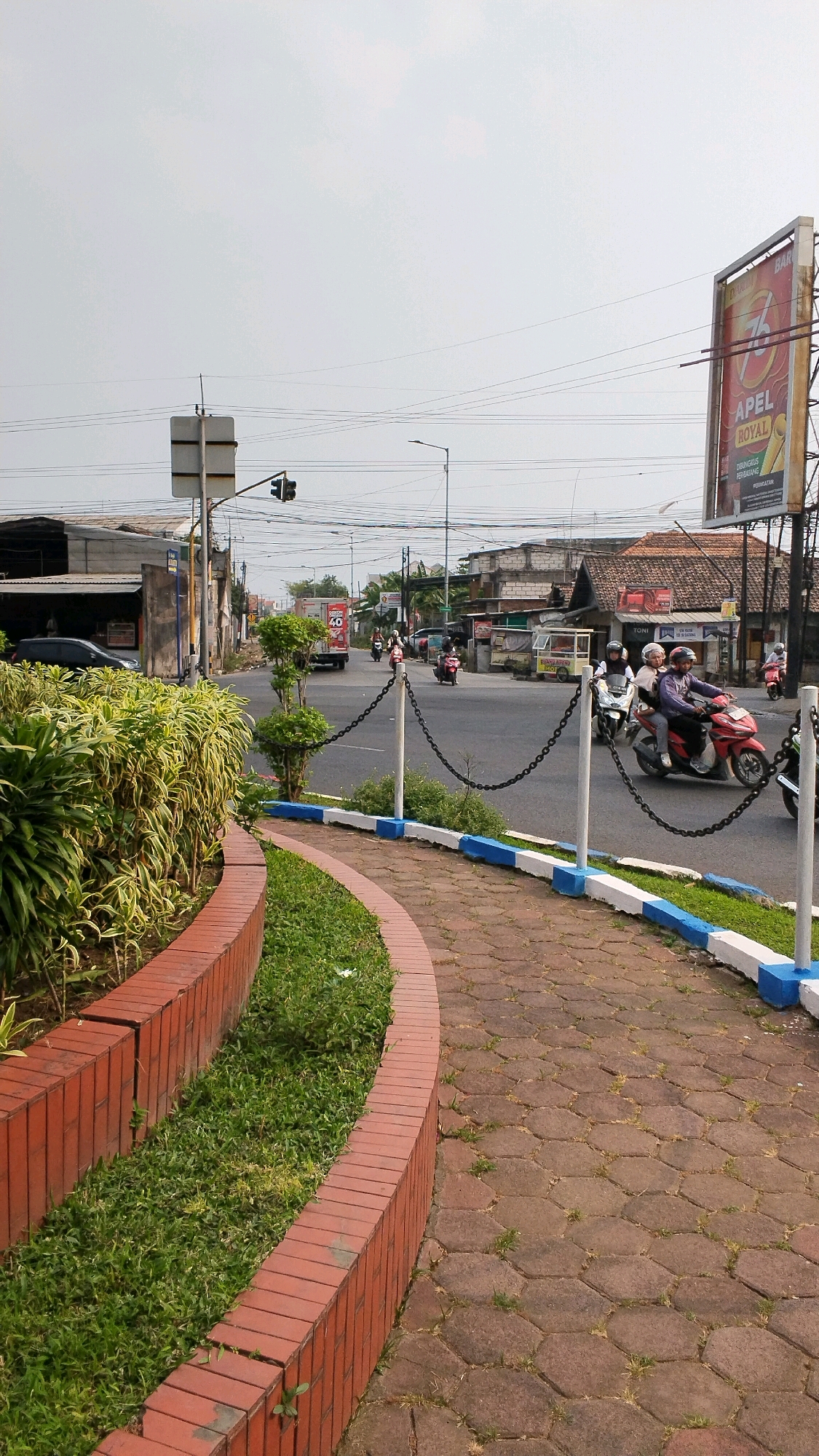 This is a ground-level, eye-level, vertical, medium shot of a street intersection with a sidewalk and garden area in the foreground. The scene is set in Banjaragung, Indonesia. The sky is overcast and light.
In the foreground, a curved brick planter box filled with lush green grass and some leafy green plants lines the left side of the image. To the right of the planter is a hexagonal paver sidewalk, also curving into the distance. The sidewalk is bordered by a low curb painted with alternating blue and white sections. A chain-link fence with white posts separates the sidewalk from the street.
The street is paved and appears to be a moderately busy intersection. Several motorcycles are visible, with riders on them, some turning and others proceeding straight. One red motorcycle with two riders is prominent in the mid-right of the frame. A white truck with "40" written on its side is heading away from the camera in the distance. Various buildings with tiled roofs and weathered facades line the streets. A large billboard with text and an image is visible on the right side of the street. Traffic signals are present at the intersection. Power lines crisscross the sky above the street. The overall impression is a typical urban street scene in Indonesia.
