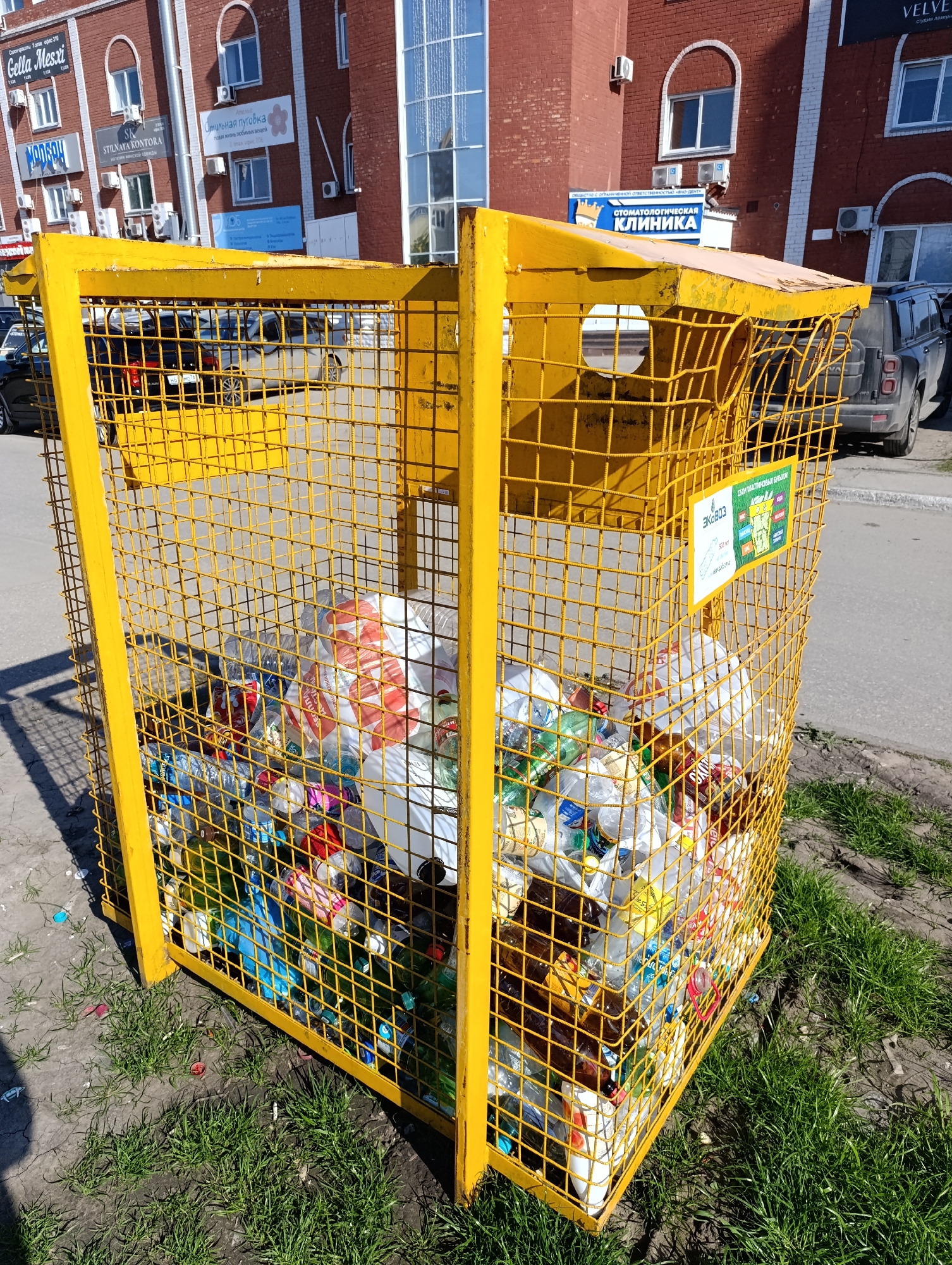 The image shows a yellow metal cage-like container overflowing with plastic bottles and bags, likely for recycling. The container is located outdoors on a paved surface with some grass patches visible. In the background, there are multi-story buildings with brick facades, windows, and signs in Russian, indicating shops and services such as a beauty salon (Gella Mesxi), a women's clothing store (Stilnaya Kontora), and a dental clinic. Cars are parked on the street beside the container.