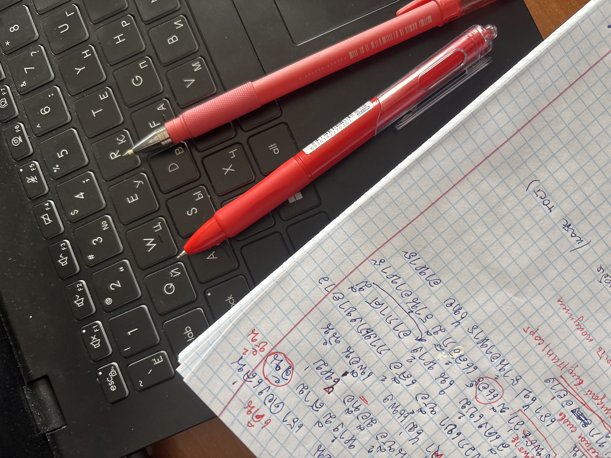 The image presents a close-up view of a workspace, likely indoors, focused on a black laptop keyboard. Resting on the keyboard are two red pens and a sheet of graph paper filled with handwritten notes in an unidentified script, possibly Thai, and what appears to be some Russian phrases. The notes contain a mix of text and circled symbols. The scene conveys an environment of study or note-taking. The location is not indicated within the visual context.