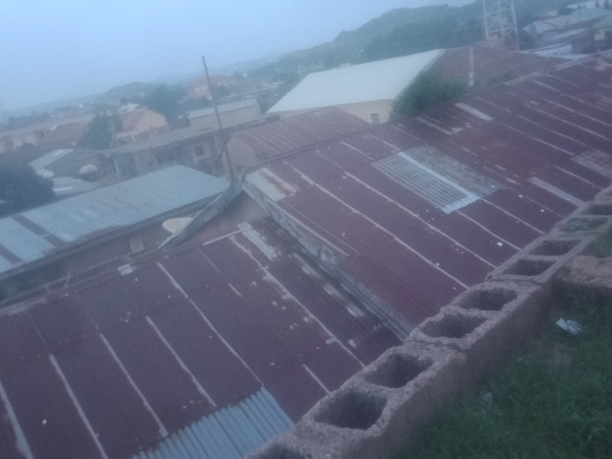 A high-angle, wide shot captures a hazy view of a dense urban landscape under an overcast sky. The foreground is dominated by the weathered, reddish-brown corrugated metal roofs of several buildings, interspersed with sections of lighter, grey corrugated metal. The roofs are uneven and show signs of age and wear. A rough, grey concrete cinder block wall runs along the right edge of the frame, with patches of green grass visible below it. In the mid-ground, a white-roofed building stands out, with a yellow-walled building adjacent to it. Further into the distance, more buildings and trees are visible, all softened by the atmospheric haze. A faint silhouette of a hill or mountain range can be seen in the far background, adding to the sense of depth and distance. The overall impression is of a somewhat dilapidated urban environment, likely in a region experiencing poor air quality or fog.