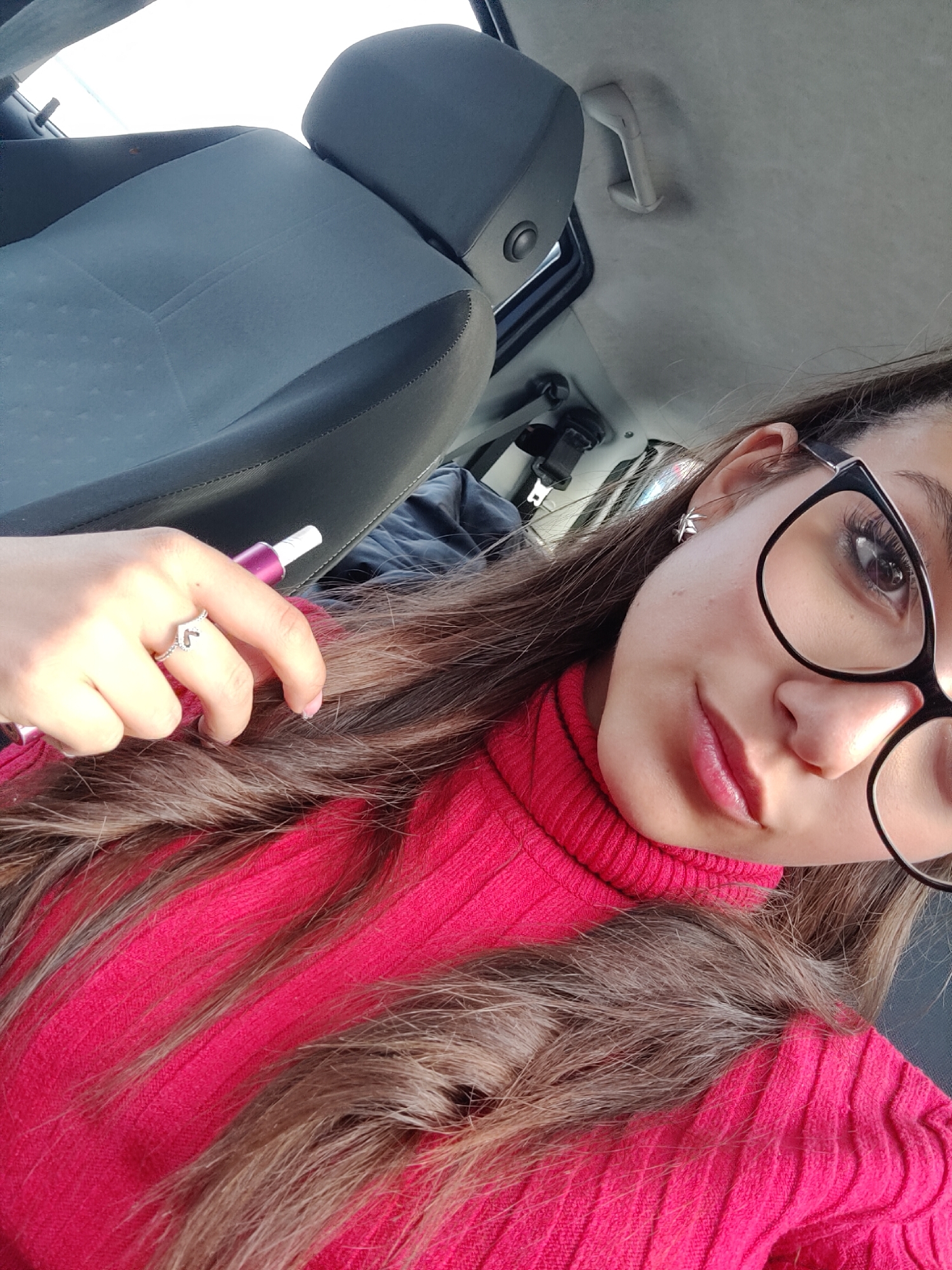 The image shows a woman inside a car, likely in the driver's seat, given the angle and proximity to the steering wheel. The interior of the car is visible, with a gray seat and the car's ceiling. The woman is wearing a red turtleneck sweater and glasses. She is holding a purple and white electronic cigarette in her left hand. A silver ring is visible on her finger.
There is not enough information to conclude that the location is in the city of Plovdiv, Bulgaria.