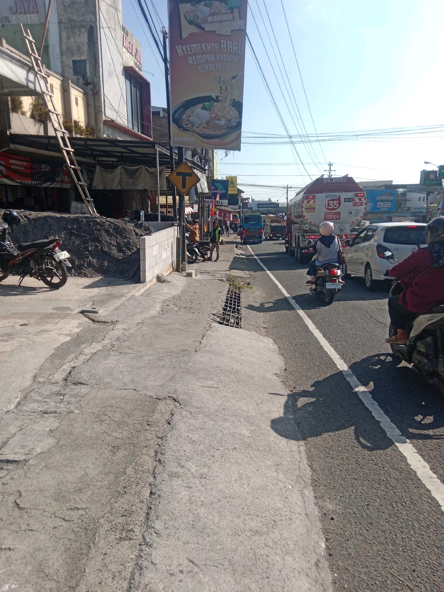 This is a photograph of a street scene in Kertek, Indonesia. The image depicts a busy street with moderate traffic, including motorcycles and cars. In the foreground, a concrete sidewalk is cracked and uneven, with a small pile of sand to the left. A motorcycle is parked on the sidewalk. Further down the street, a fuel tanker truck is visible, along with several other vehicles. A pedestrian wearing a reflective vest is standing on the side of the road. Buildings line the street, some with signs advertising businesses. Overhead, power lines crisscross the sky. The weather appears to be sunny.