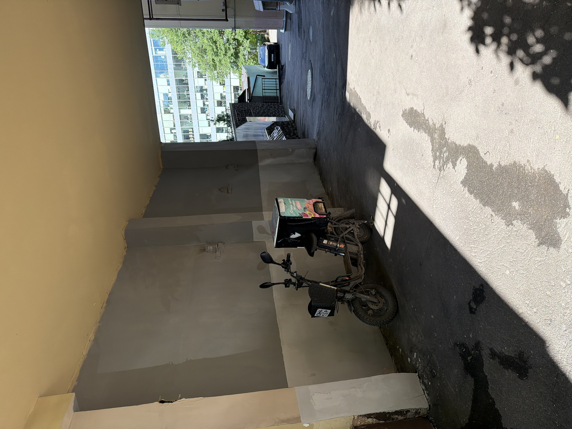 The image shows a narrow alleyway in Moscow, Russia, on a sunny day. On the left side of the alley, a dark-colored electric scooter with a large delivery box on the back is parked. The scooter has a basket in the front with a license plate that reads "OH4 15P". The alley walls are a light gray color, with some patches of white paint. Sunlight streams into the alley, casting shadows on the ground. In the background, a modern office building with many windows is visible, along with some green trees. A black car is parked further down the alley. The ground of the alley is paved with asphalt, and there are some wet patches on the ground.