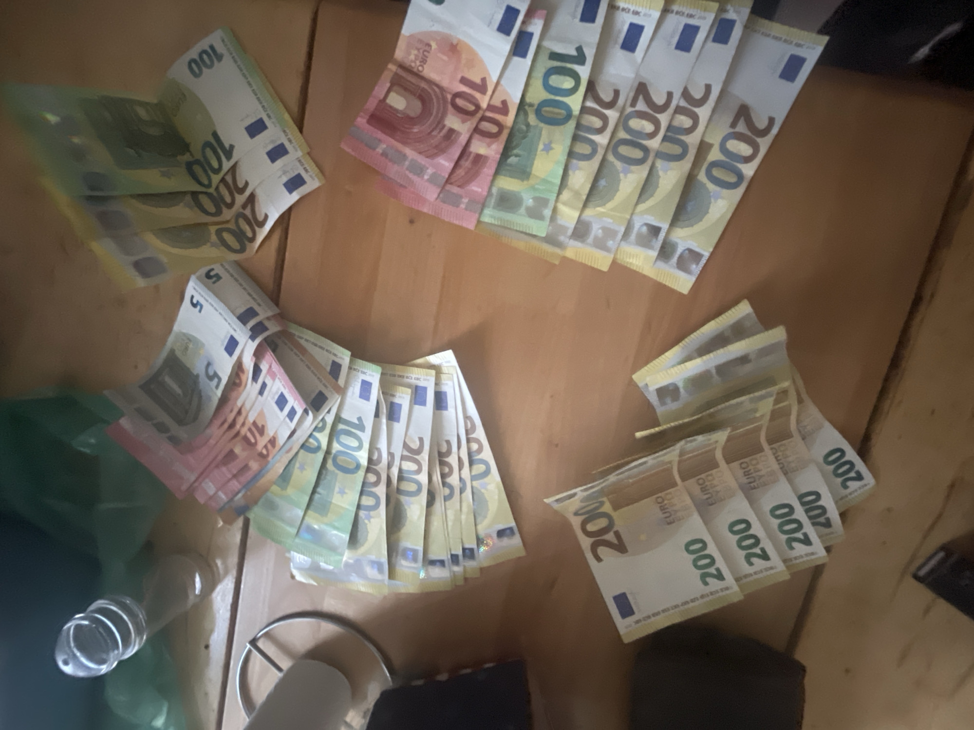 An overhead shot shows a variety of euro banknotes scattered across a wooden table. The bills are predominantly of denominations 200, 100, and 10 euros. Some bills are fanned out, while others are stacked. A clear plastic bottle with a coiled neck and a small metal ring is visible in the upper left corner, next to a crumpled green plastic bag. To the left of the banknotes is a white cylindrical object with a metal ring surrounding its base, positioned on a dark rectangular object. The scene is lit from above, creating subtle shadows on the wooden surface.