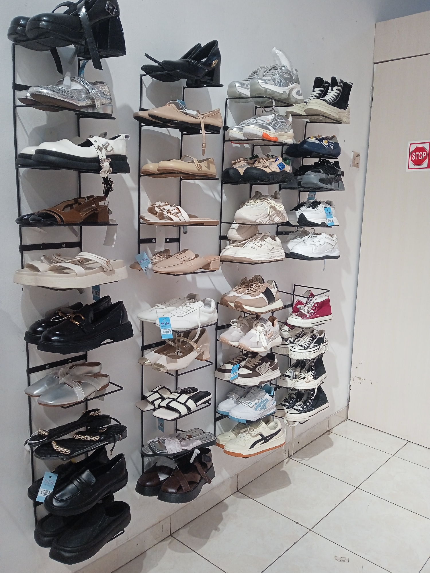 The image depicts a shoe store interior, likely located in Semarang, Indonesia, as evidenced by the "STOP" sign written in Indonesian. The scene features multiple wall-mounted shelves displaying a wide variety of women's and girls' footwear, including heels, sandals, sneakers, and flats. The shoes are organized by style and color, with some still bearing price tags. The lighting suggests an indoor commercial setting, with the floor tiled in a neutral color. The overall impression is one of a well-stocked retail environment dedicated to footwear.