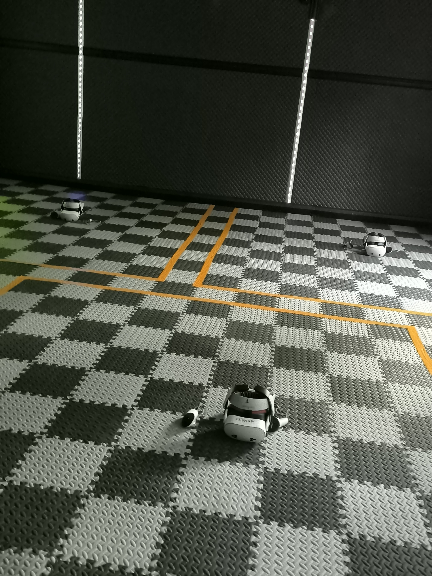 The image depicts an indoor virtual reality (VR) gaming or training space located in Trastenik, Bulgaria. The scene showcases a dark, possibly padded or sound-insulated wall in the background, creating a focused environment. The floor is covered with interlocking foam mats forming a distinct checkerboard pattern of alternating dark grey and light grey tiles. Bright, vertical LED light strips are mounted on the walls, providing illumination.
Three white virtual reality headsets, identifiable as Meta Quest devices, are strategically placed on the floor, each accompanied by a controller. The headset in the foreground prominently displays "QUEST" and a numerical identifier, possibly "65700CM," on its side. Orange tape is meticulously laid out across the checkerboard floor, delineating play areas or boundaries within the space.
No individuals are present in the image, suggesting the area is either prepared for an upcoming session or has just been used. The arrangement of the equipment and the structured layout of the room indicate a dedicated and organized facility for VR experiences. The artificial lighting and lack of external elements make the time of day and weather irrelevant to the indoor setting.