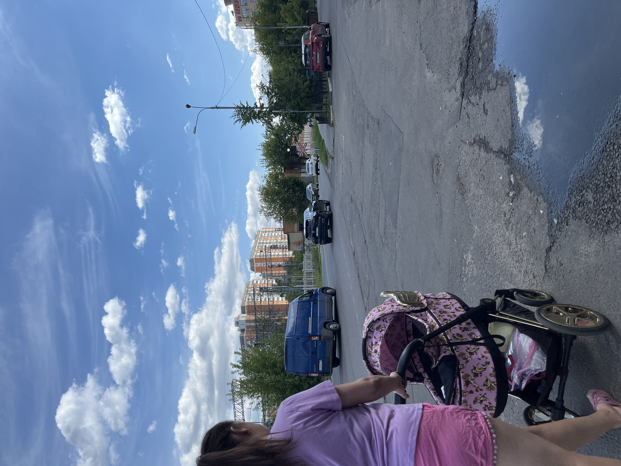 In Balashikha, Russia, on a bright, cloudy day, a woman from behind pushes a pink baby stroller with a gold trim and a cherub pattern along a cracked asphalt road. She wears a light purple t-shirt and pink shorts with a pom-pom trim. A blue van drives away from the camera on the asphalt road. Several cars are visible further down the road, including a black SUV and a red SUV. In the background, residential buildings with orange facades stand amidst green trees. A streetlamp is visible on the right side of the frame.