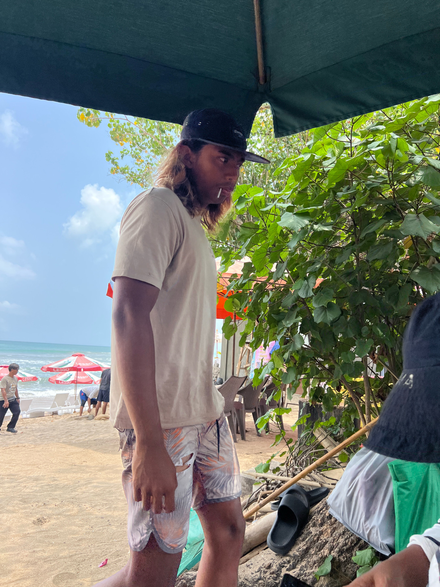 A young man with long, light brown hair and a black baseball cap is standing on a sandy beach. He is wearing a light-colored t-shirt and patterned shorts. He is holding a white object, possibly a cigarette or lollipop, in his mouth. In the background, the ocean is visible with waves crashing onto the shore. Several red and white umbrellas are set up on the beach, along with lounge chairs. Other people are visible in the background, some walking and others appearing to be working. Lush green foliage and tree branches are on the right side of the image, partially obscuring the view. A dark green umbrella is overhead. The overall setting suggests a sunny day at a beach resort in Kuta, Indonesia.