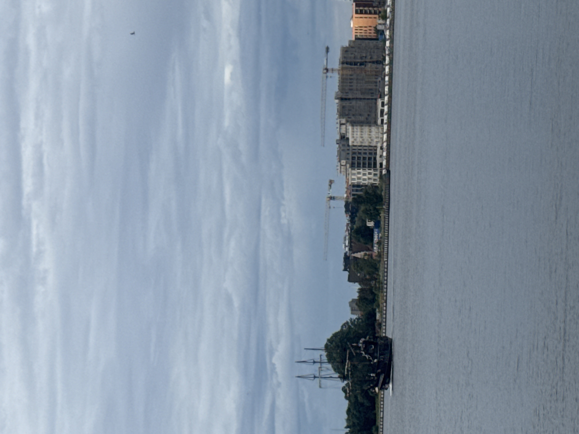 In the city of Gdańsk, Poland, a pirate-themed ship sails on a wide body of water under a cloudy sky. The ship, black with multiple masts and rigging, is positioned on the left side of the frame, leaving a wake behind it. In the background, along the shoreline, construction cranes are visible above a line of buildings, some of which appear to be under construction. Trees are interspersed with the buildings, and a thin line of what seems to be a breakwater or embankment runs along the water's edge. The water itself is calm and reflects the overcast sky. A single bird is visible flying in the upper right portion of the sky.