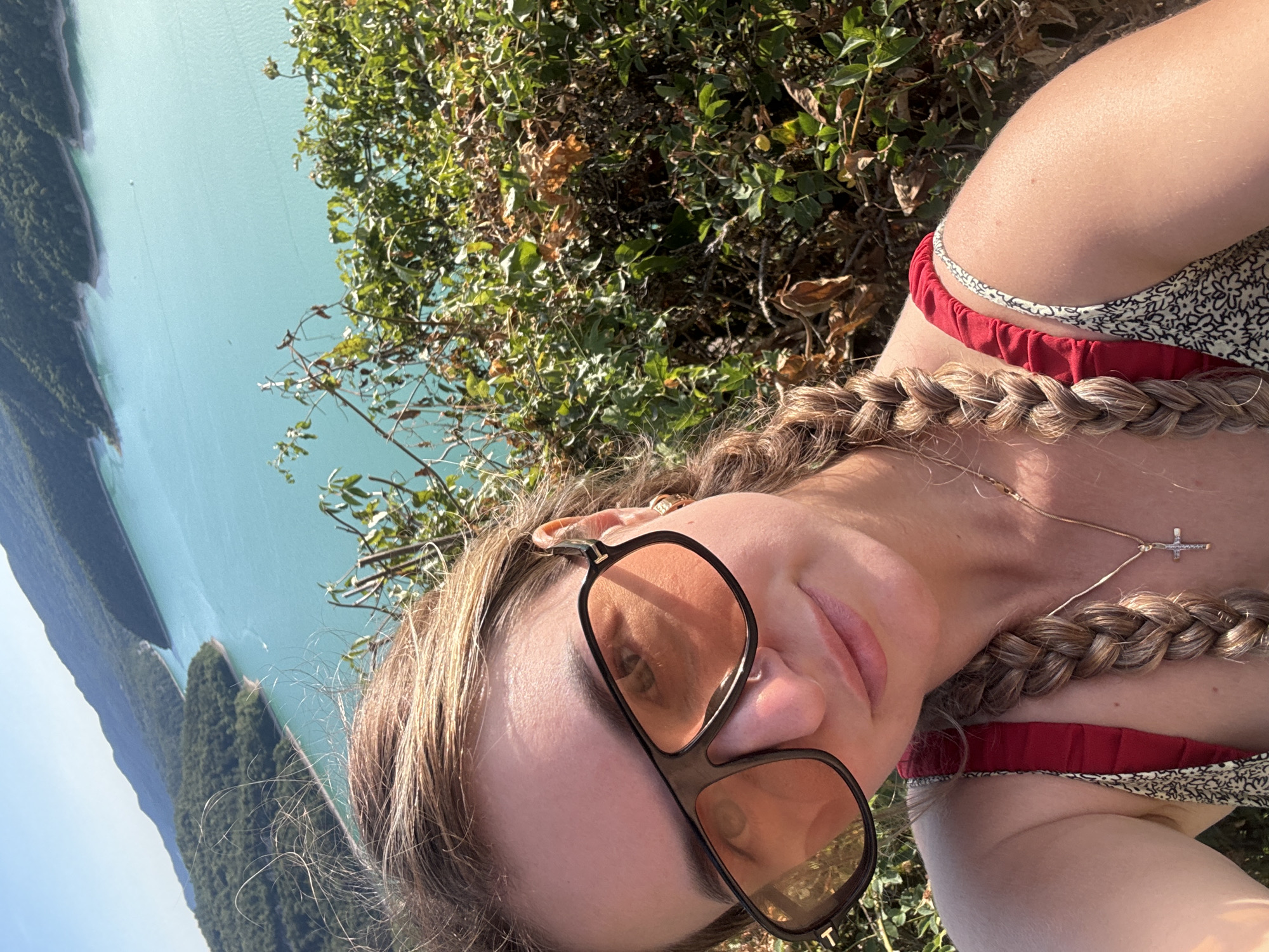 A young woman with blonde hair styled in two braids is taking a selfie outdoors, with a vibrant turquoise lake and tree-covered mountains in the background. She is wearing large, dark-rimmed sunglasses with orange lenses, a red and white patterned top with red ruffled straps, and a gold necklace with a cross pendant. The location appears to be the Lac de Vouglans, near the town of Moirans-en-Montagne, France, a popular tourist destination known for its scenic beauty and recreational activities. The sun is shining brightly, casting shadows and highlighting the lush green foliage around her.