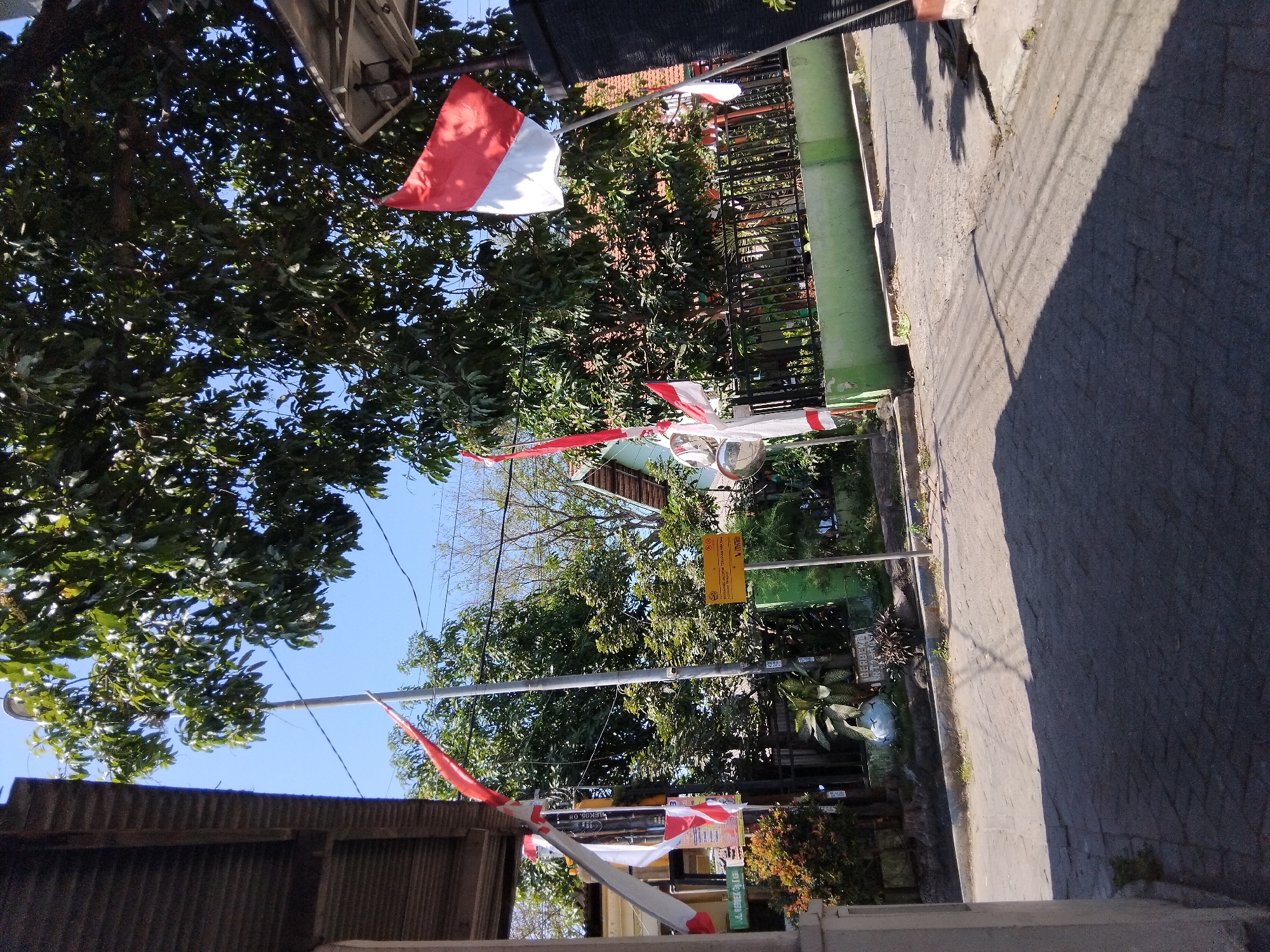 In the city of Berbek, Indonesia, a street scene unfolds under a clear blue sky. The foreground features a paved road, with shadows cast by overhead trees and structures. On the left, a portion of a building with a corrugated roof is visible. Several Indonesian flags, red and white, are prominently displayed on poles along the street. One flag flies high, while others are seen at varying heights and angles. Behind the flags, a green fence encloses a property with trees and a building with a reddish-brown roof. A street sign, yellow with black text, is mounted on a pole. The overall atmosphere suggests a public area decorated for a national occasion.