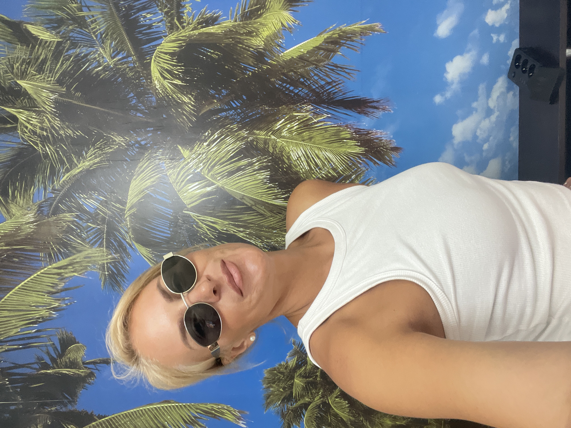 A blonde woman wearing sunglasses and a white tank top takes a selfie against a backdrop of palm trees and a blue sky with clouds. She is looking towards the camera with a slight smile. A black remote control rests on a dark surface in the lower right corner of the image. The scene appears to be indoors, with the palm tree and sky elements likely being a mural or wallpaper. There is no indication of the city of Gagra, Abkhazia.