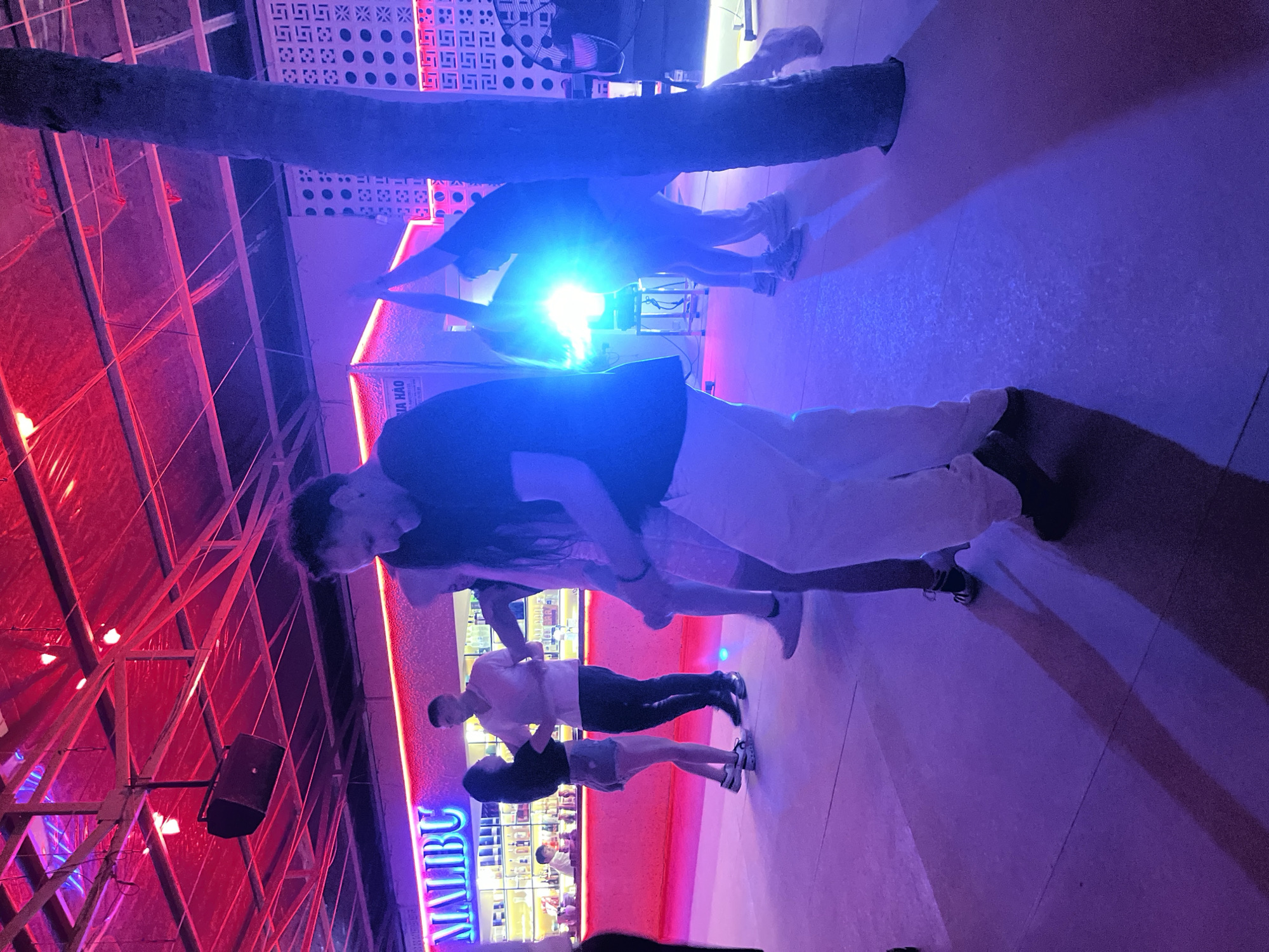 The image captures a lively nighttime scene at a bar or club in Da Nang, Vietnam. In the foreground, a couple is dancing closely, illuminated by purple and pink neon lights. The woman wears a black top and denim shorts, while the man is dressed in a light pink shirt and dark pants. Behind them, another couple is also dancing, with the woman in a black top and shorts and the man in a black t-shirt and grey sweatpants.
The backdrop is a vibrant bar area with a long counter filled with bottles, bathed in pink neon light. The word "MALIBU" is prominently displayed in neon blue lettering on the counter. Above, a tree trunk extends into the frame, possibly indicating an outdoor or semi-outdoor seating area. The ceiling features a complex structure with metal beams and red translucent material, suggesting a modern, somewhat industrial design.
The lighting is predominantly purple and pink, creating a party atmosphere. The bright light source in the mid-ground suggests a stage or spotlight, possibly from a DJ or live performance. The overall mood is energetic and festive, with the dancers appearing to be enjoying themselves. The image is a snapshot of a social gathering, likely a weekend night out in Da Nang.