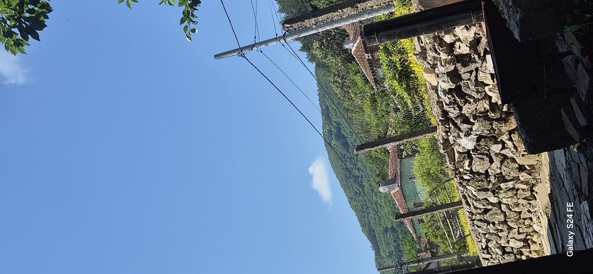 The image captures a rural scene in Malko Tarnovo, Bulgaria, characterized by a stone wall in the foreground, a wire fence, and houses nestled against a lush, green hillside. The sky is a clear, vibrant blue with a solitary white cloud. A power pole with wires stretches across the frame. The stone wall is constructed from rough, irregular rocks. Behind the fence, traditional houses with red-tiled roofs are visible, surrounded by dense vegetation and trees. The overall atmosphere is tranquil and suggests a warm, sunny day in a mountainous region.