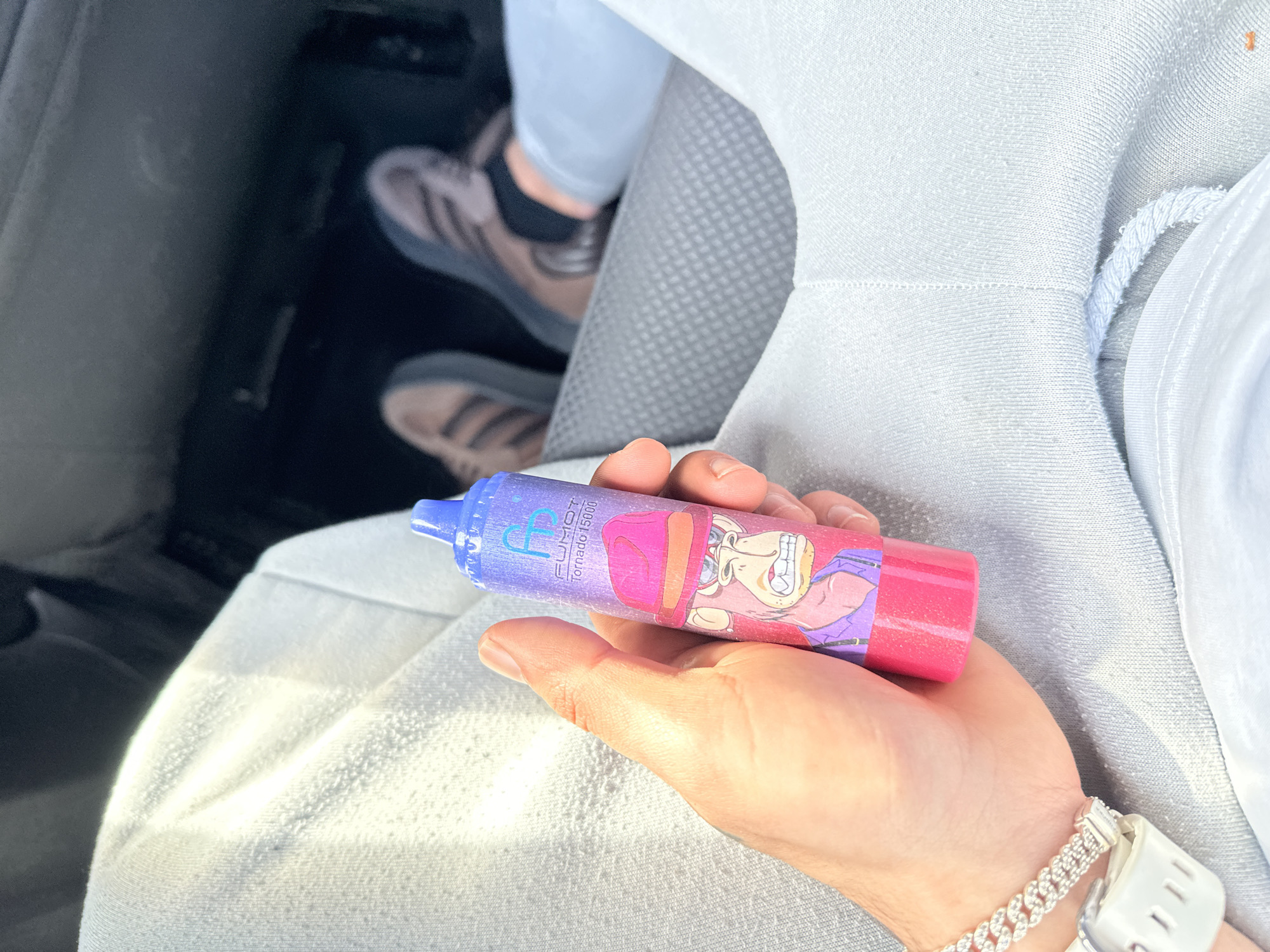 A person is seen holding a FUMOT Tornado 15000 disposable vape in their hand, inside a vehicle. The vape is pink and purple and features a cartoon character on its side. The person is wearing a white sweatshirt and a silver bracelet on their wrist. In the background, part of the car's interior is visible, including the floor, seat and the feet of another person wearing grey shoes. The location is not Zeist, Netherlands, as there is no verifiable information to confirm this location.