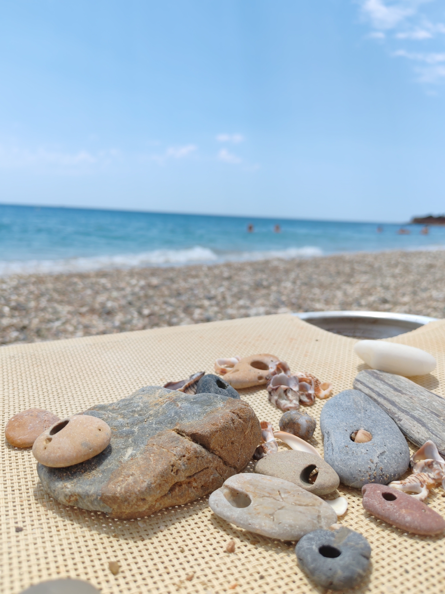 The image depicts a close-up view of various stones and seashells arranged on a textured, light-colored surface, likely a beach lounger. In the background, a pebble beach transitions to the turquoise waters of the sea, with a few distant figures visible in the water. The sky above is a clear, light blue with a few wispy clouds. The focus is on the natural objects in the foreground, which include stones of varying sizes, shapes, and colors, some with natural holes, and several small seashells. The lighting suggests it is daytime, with shadows indicating a bright sun. The overall scene evokes a tranquil beach setting. While the image is of a beach, the specific location in Makri, Greece cannot be confirmed from the visual information alone.