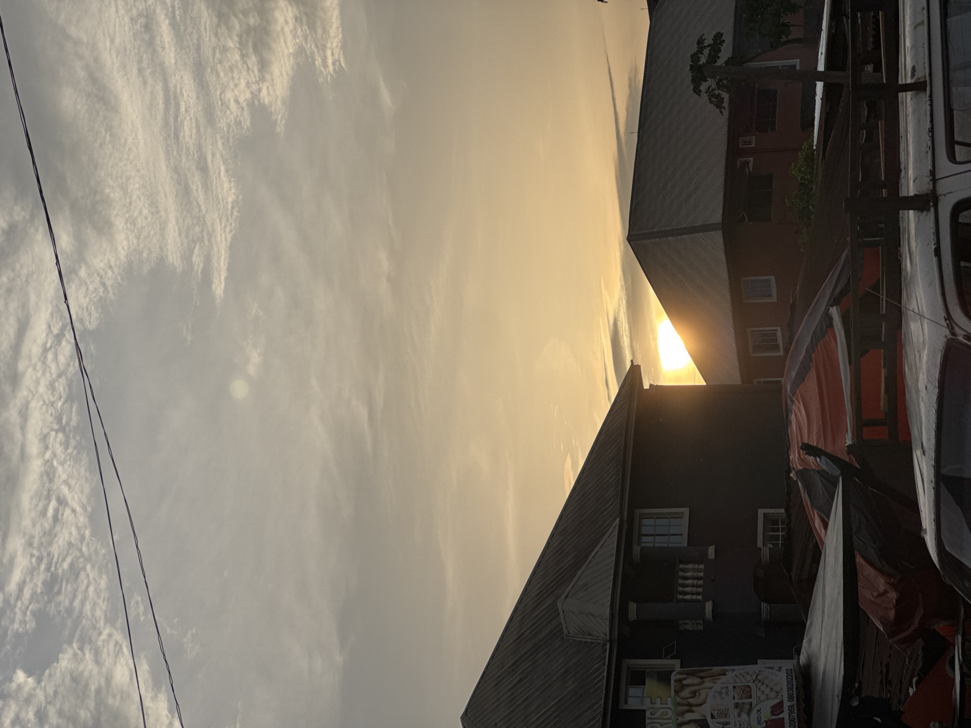The image captures a sunset view in Ihiagwa, Nigeria, framed by buildings. In the foreground, parts of a vehicle are visible on the right. The sky is filled with clouds tinged yellow by the setting sun. The building on the right is primarily red with white windows. The building on the left has a metal roof and a black facade. Utility wires are visible extending across the sky in the top left of the image.
